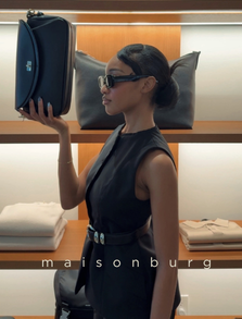 A woman in sunglasses and a sleeveless black dress holding a black bag in front of her face, standing in a store with pillows and shelves in the background.