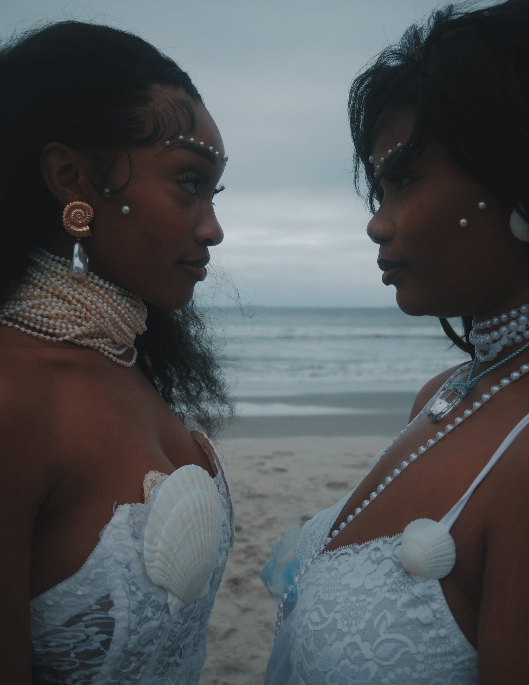 Two women standing close on a beach facing each other, wearing white lace dresses and adorned with pearl jewelry and shell accents, with ocean and cloudy sky in the background.