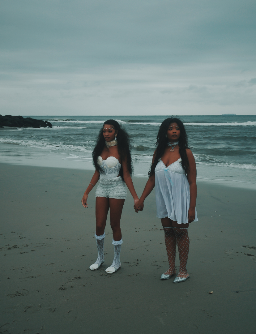 Two women in white outfits holding hands on a beach with cloudy skies and ocean waves in the background.