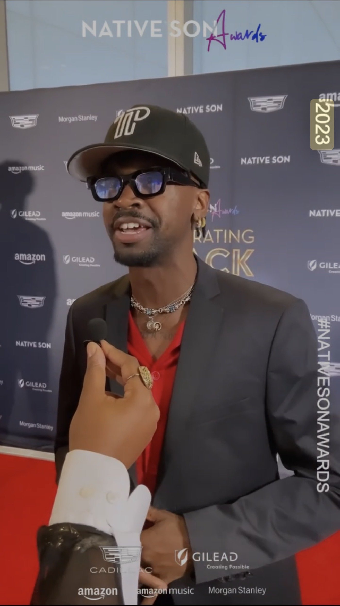 A man in a black suit and red shirt being interviewed at an event with a backdrop that says 'NATIVE SON Awards' and features logos of Morgan Stanley, Amazon Music, Gilead, Cadillac, and other sponsors.