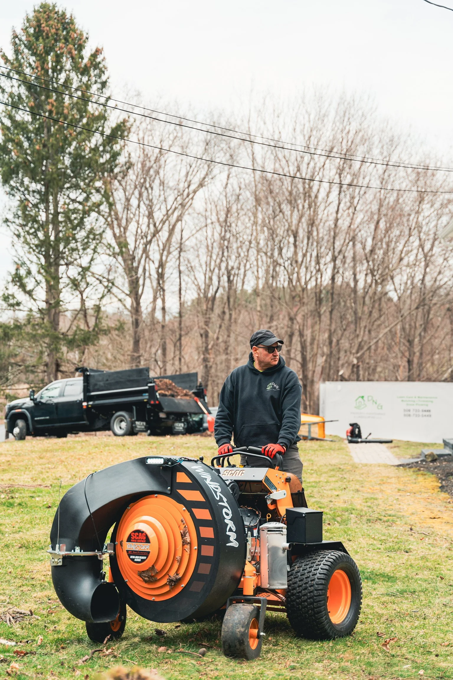 Landscaping worker operating an orange and black leaf vacuum in a grassy yard, with a utility truck and leaf-filled trailer behind him.