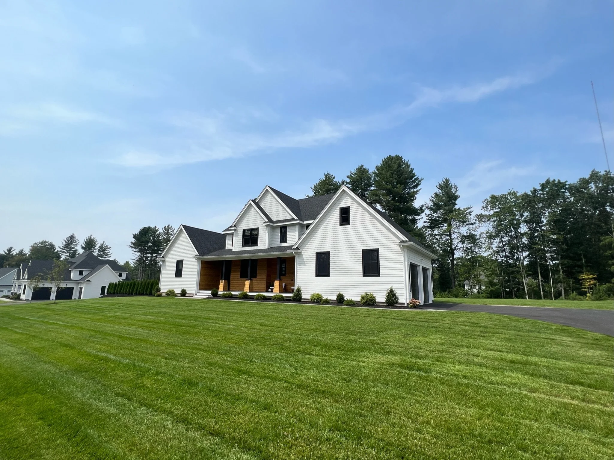 A large white house with black roofing, front porch, and multiple windows, surrounded by a well-kept lawn with striped grass, trees, and a clear blue sky.