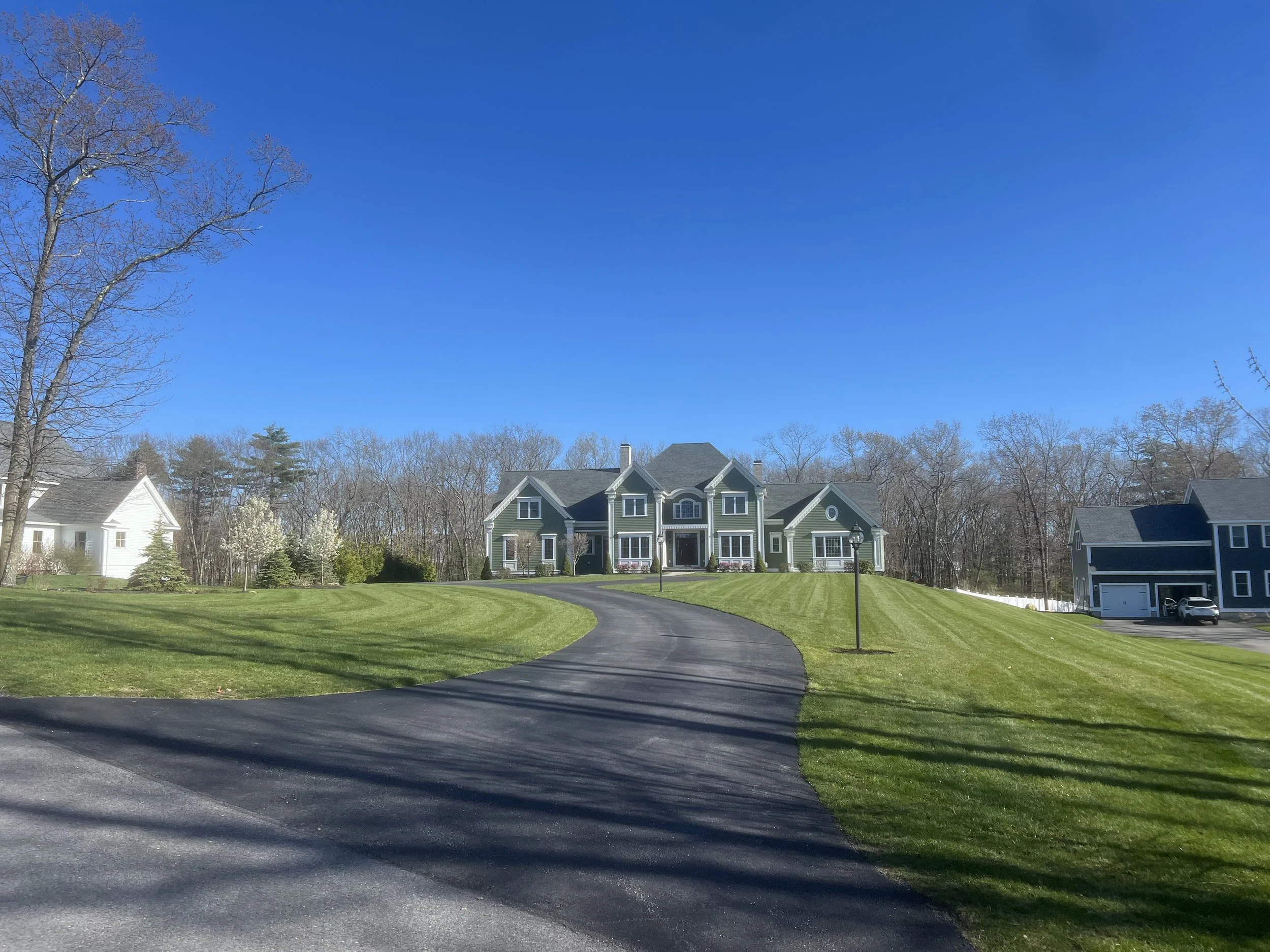 A large green house with a curved driveway surrounded by a well-manicured lawn and trees under a clear blue sky.