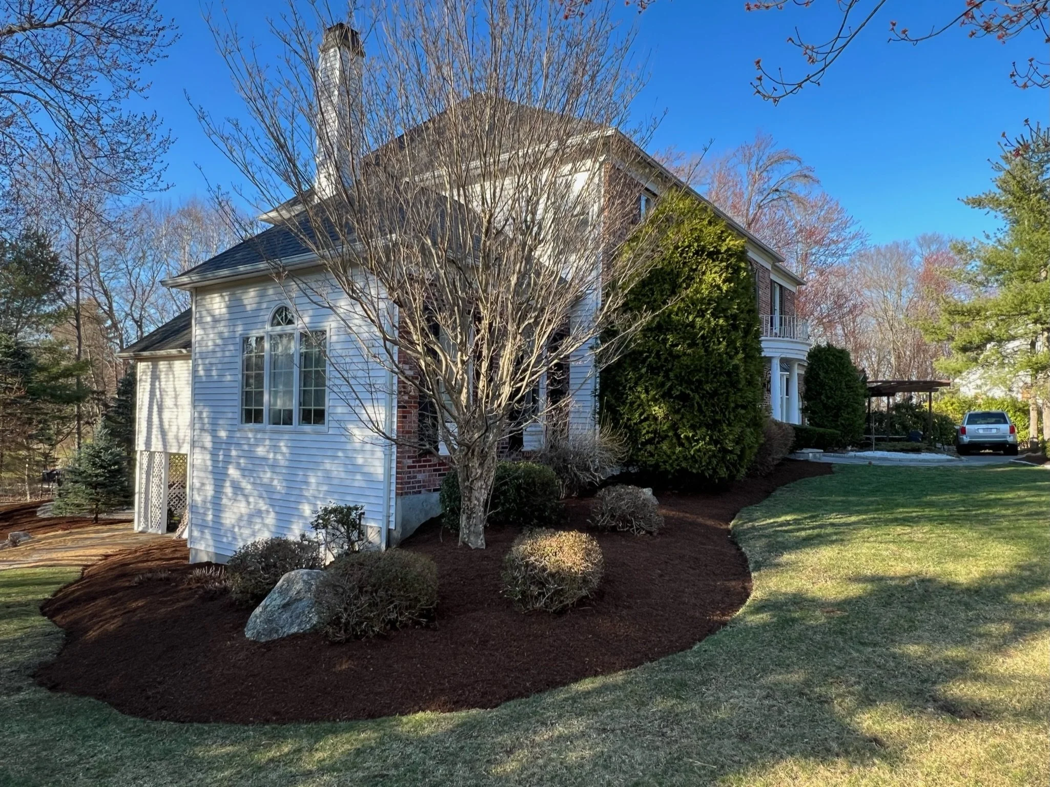 A two-story house with white siding, brick accents, and large windows, surrounded by landscaped garden beds, trees, and a lawn, with a car parked in the driveway under a carport.