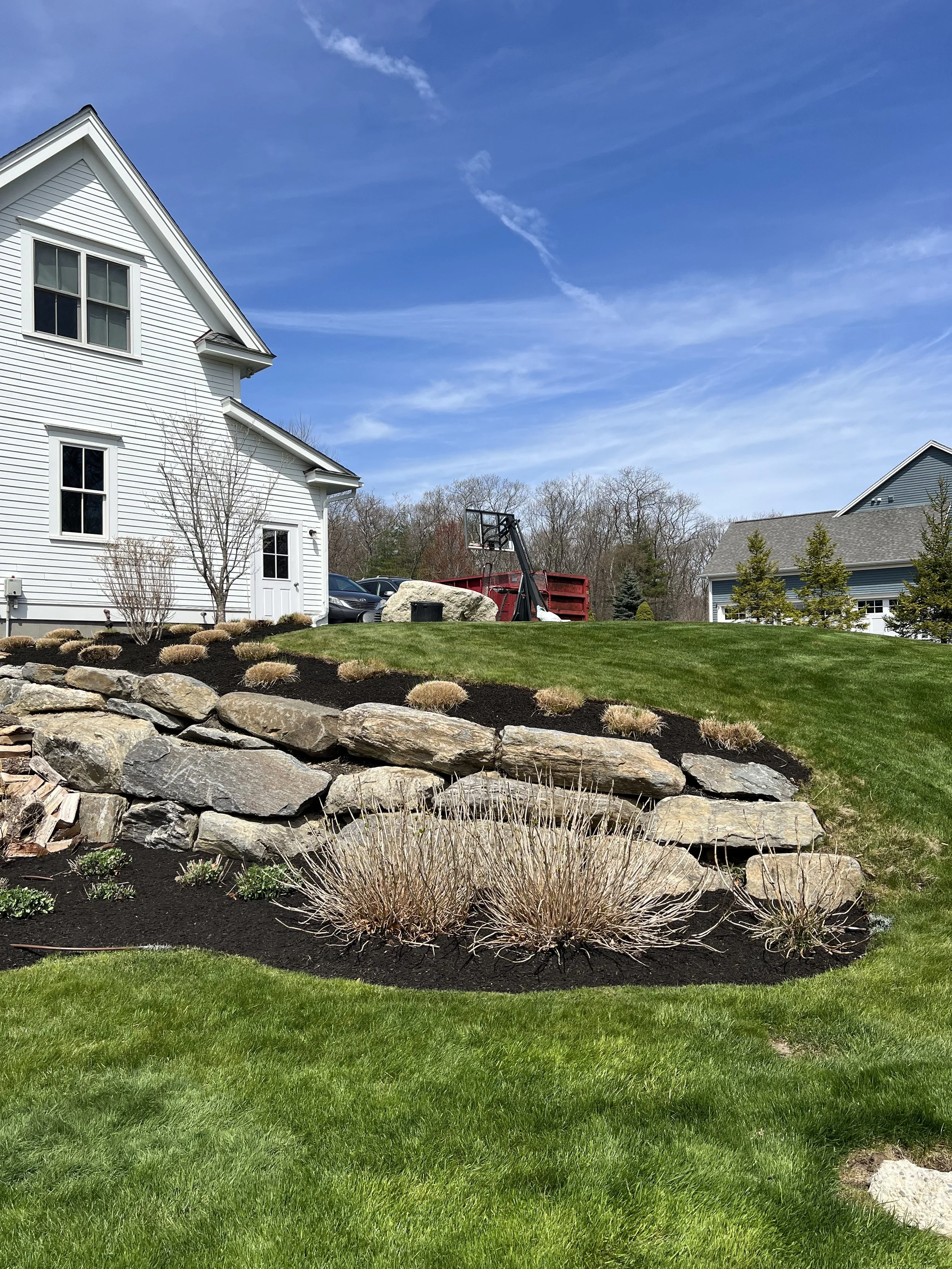  landscaped yard with green grass, large rocks, small bushy plants, and a white house with a gabled roof under a blue sky.
