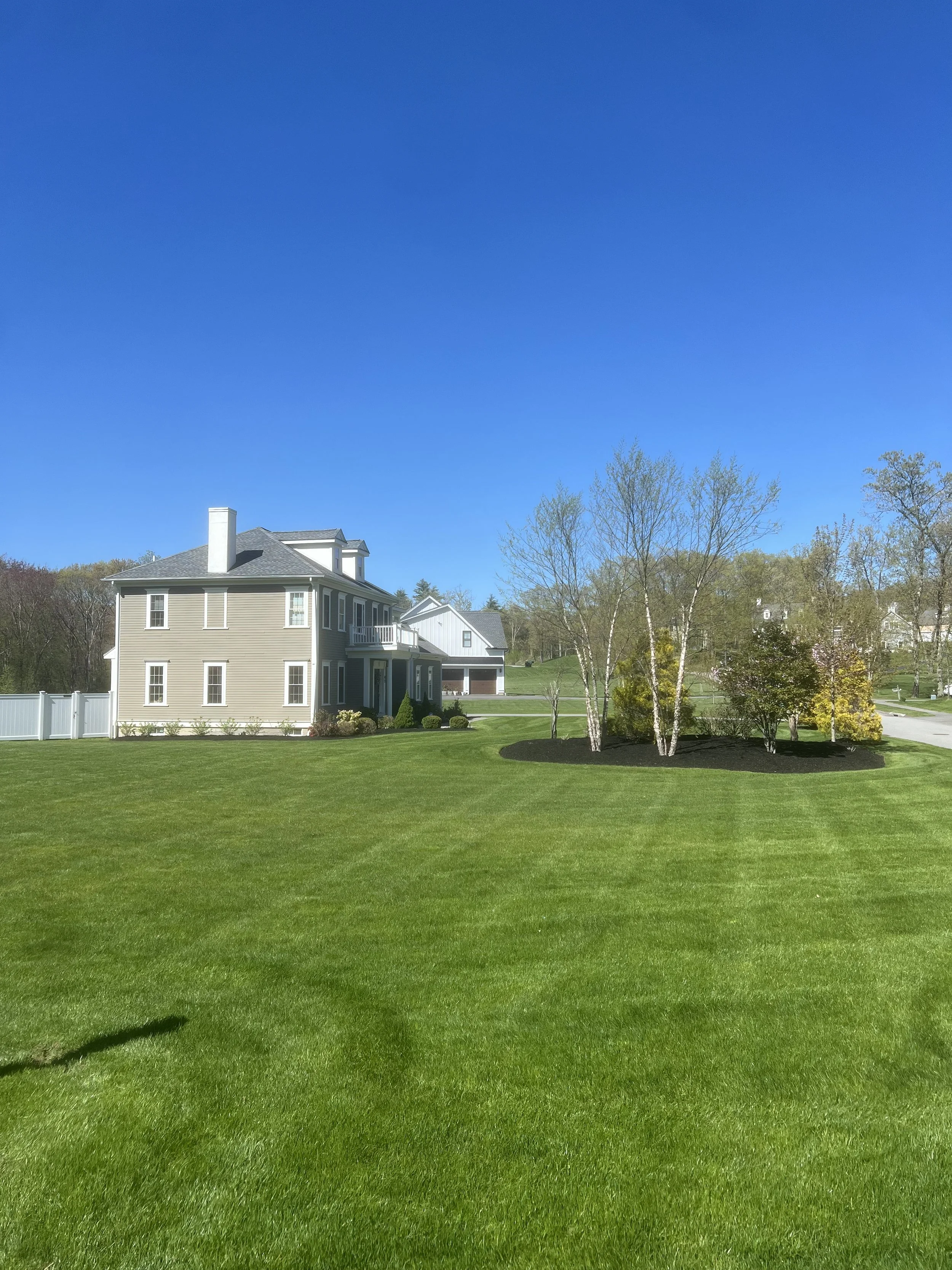 A large, beige house with white trim and a chimney, surrounded by a well-manicured green lawn and a circular flower bed with young trees and shrubs, under a clear blue sky.