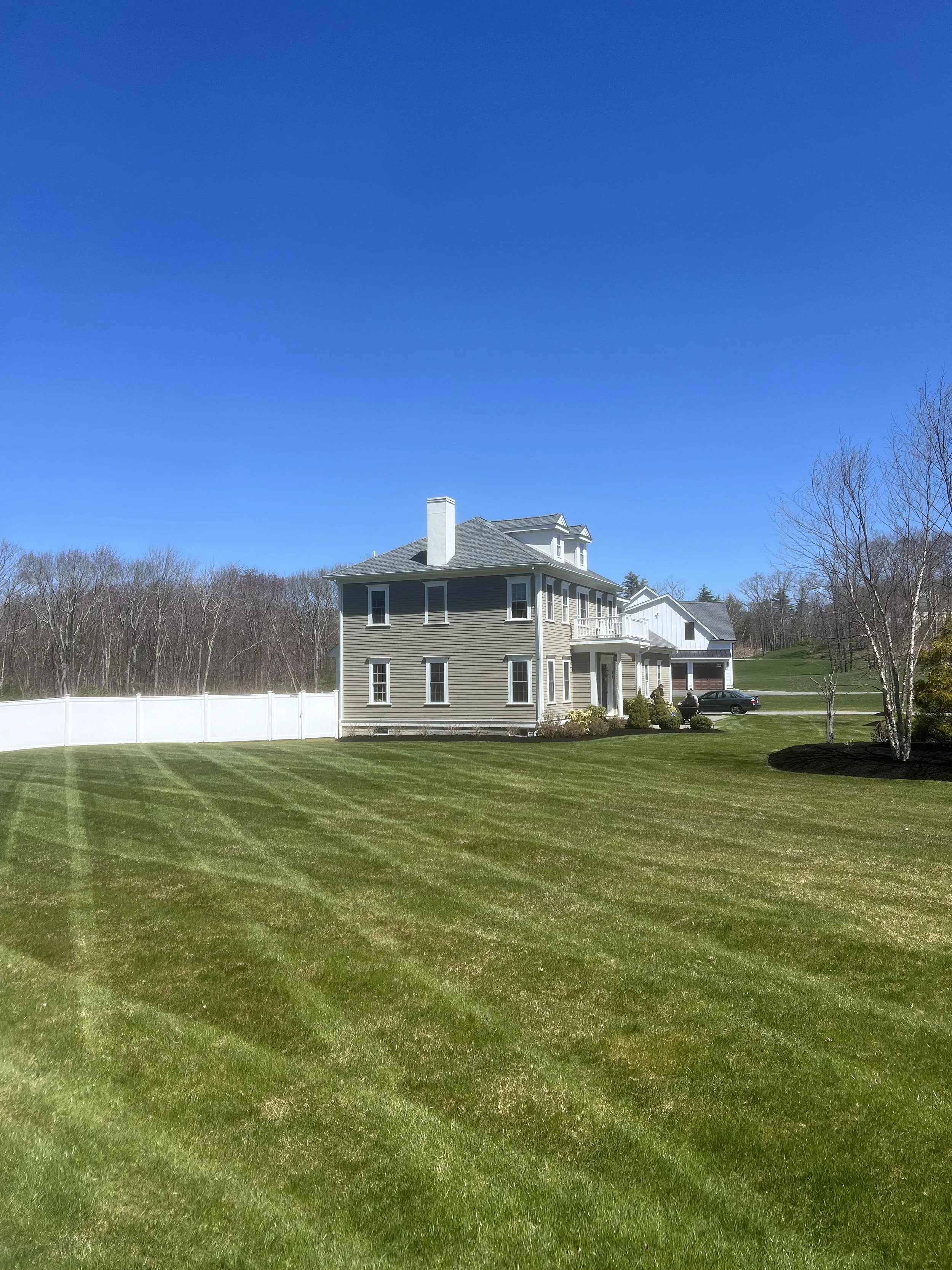 A large house with gray siding and white trim, a manicured lawn, and a white fence, under a clear blue sky.