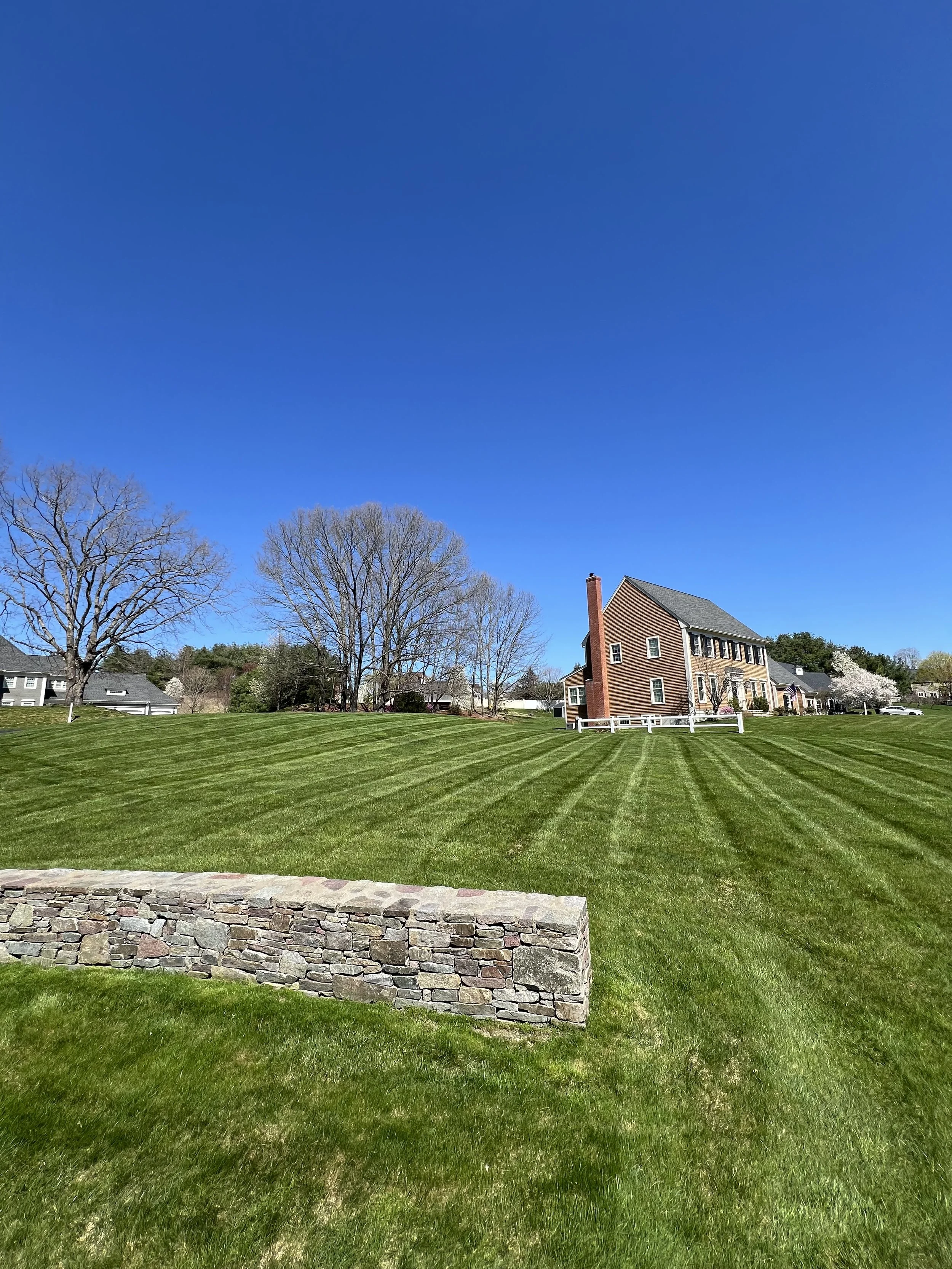 Well-maintained green lawn with stripes, a stone wall in the foreground, bare trees, and a two-story brick house with a chimney, under a clear blue sky.