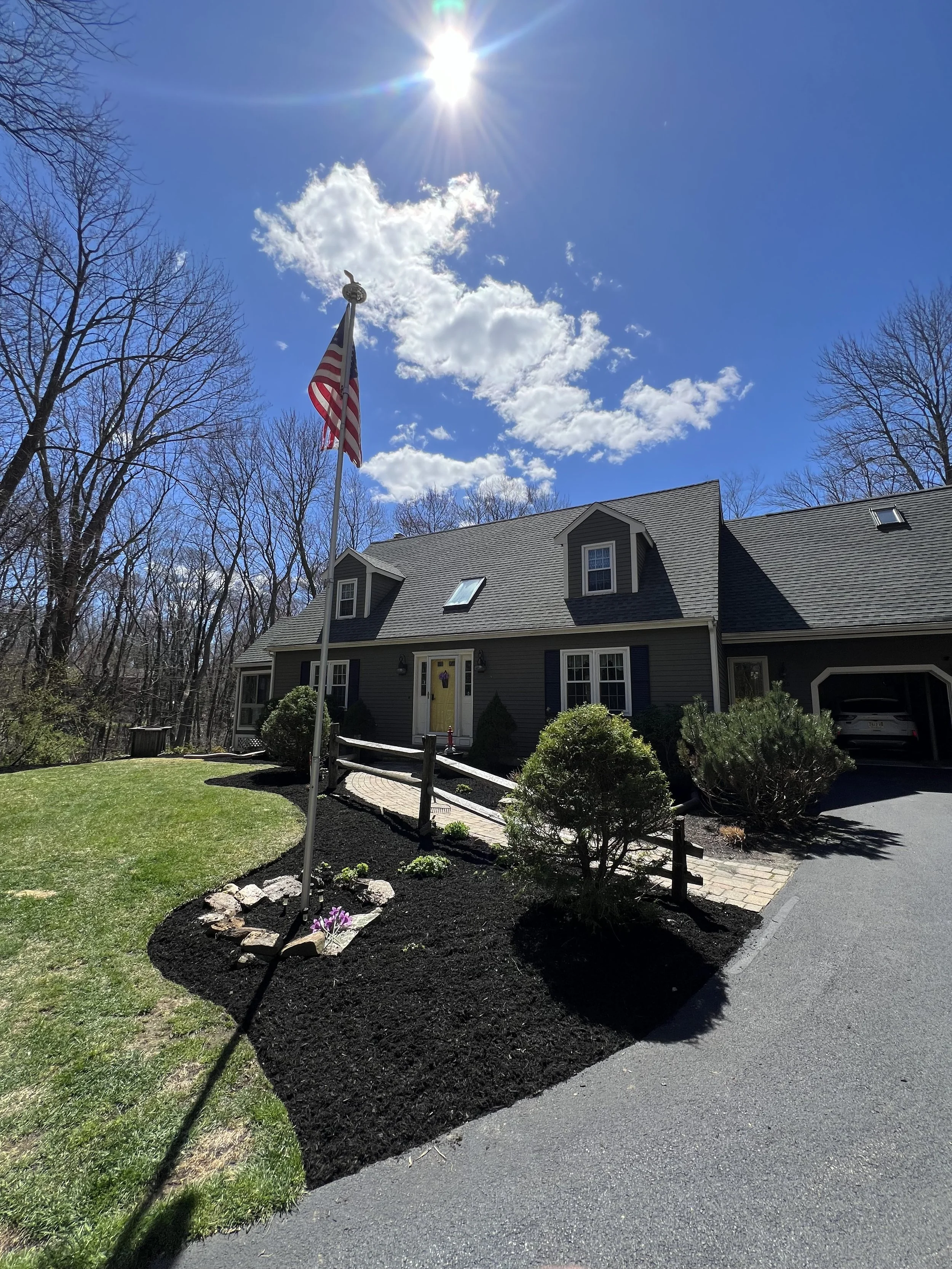 Front view of a suburban house with a dark gray exterior, yellow front door, and a landscaped yard featuring bushes and a small patch of flowers. An American flag on a pole is near the walkway, under a bright sun in a blue sky with scattered clouds.