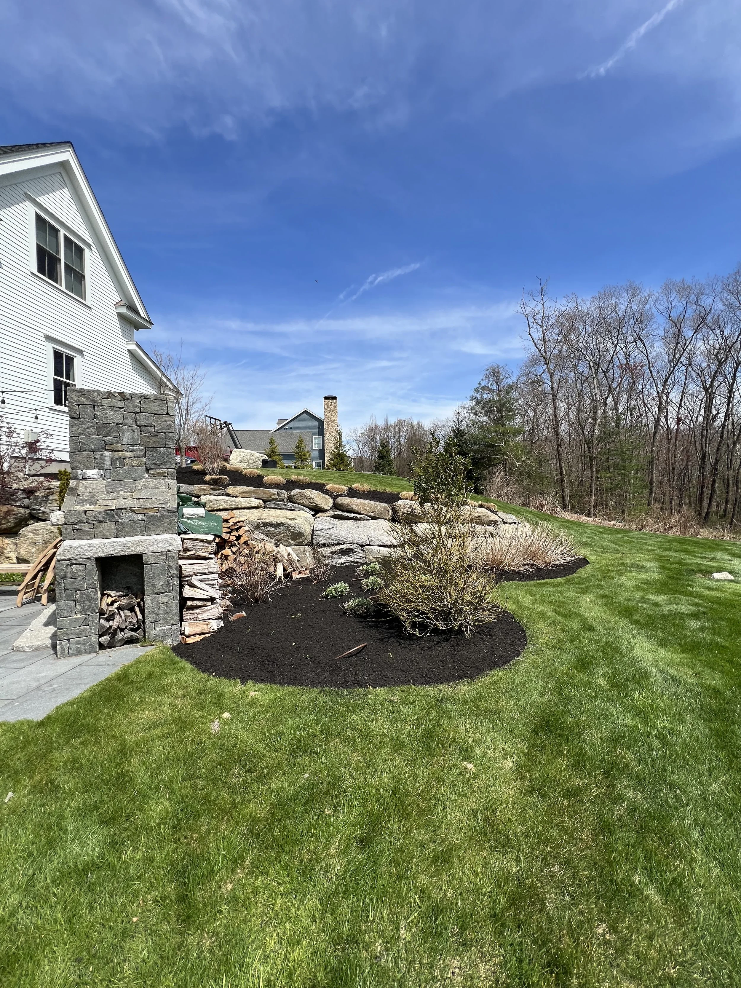 Backyard with a stone fireplace, well-maintained lawn, mulched garden beds, and a backdrop of trees under a blue sky with wispy clouds.