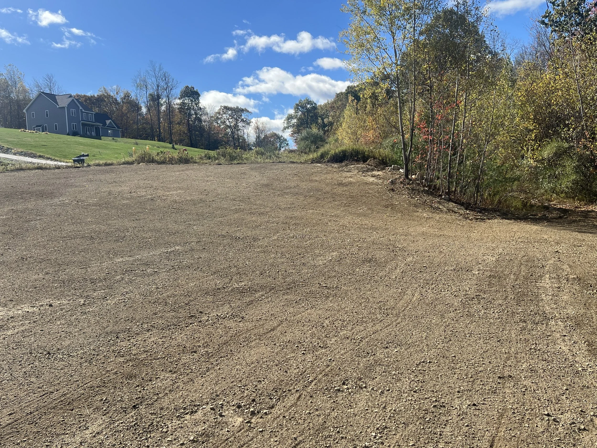 A cleared dirt lot with a house on a hill and trees in the background under a blue sky with clouds.