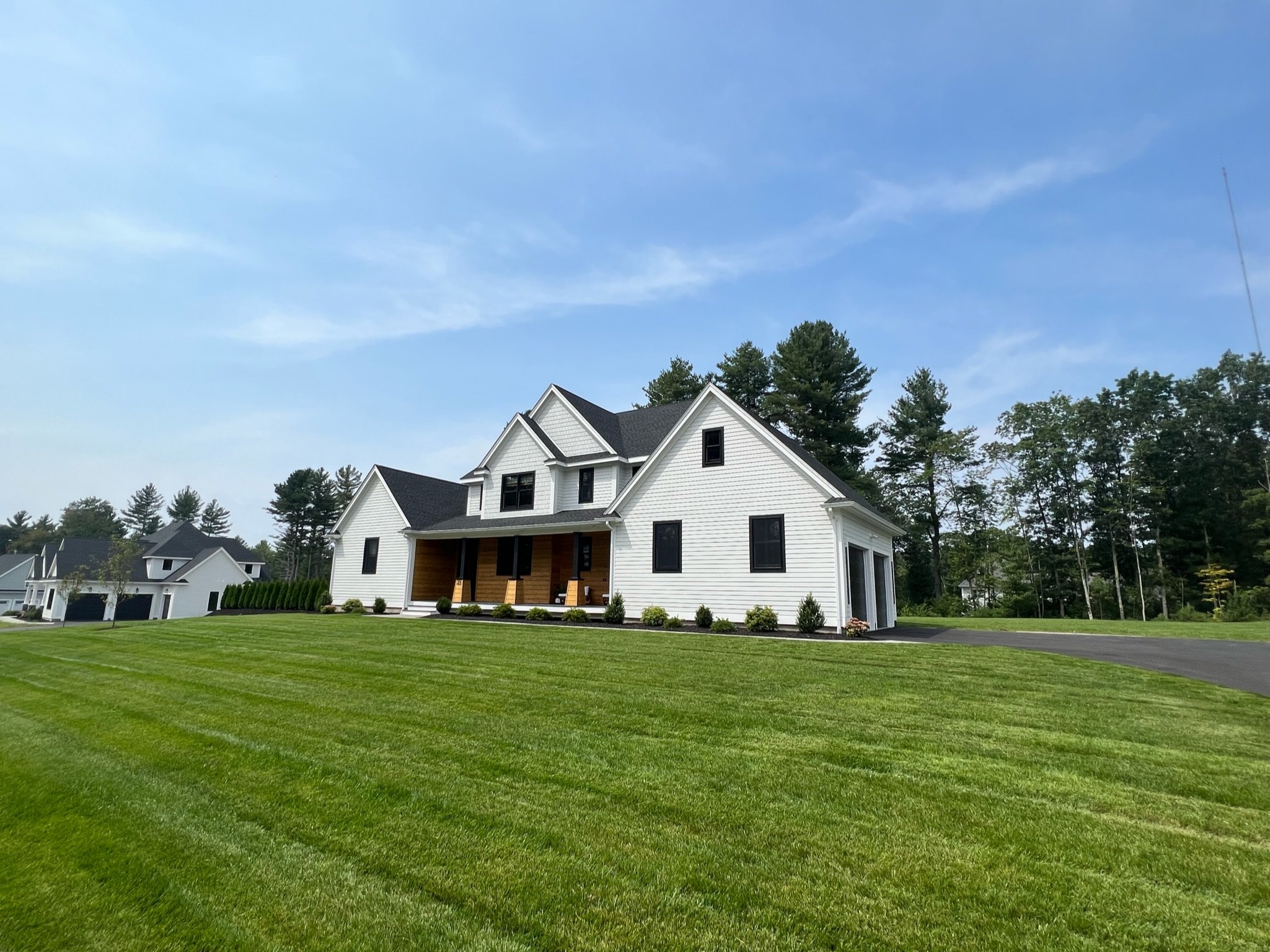 A white, two-story house with black roof and black window frames, featuring a large front porch with wooden accents, surrounded by a well-maintained green lawn and trees in the background under a blue sky.