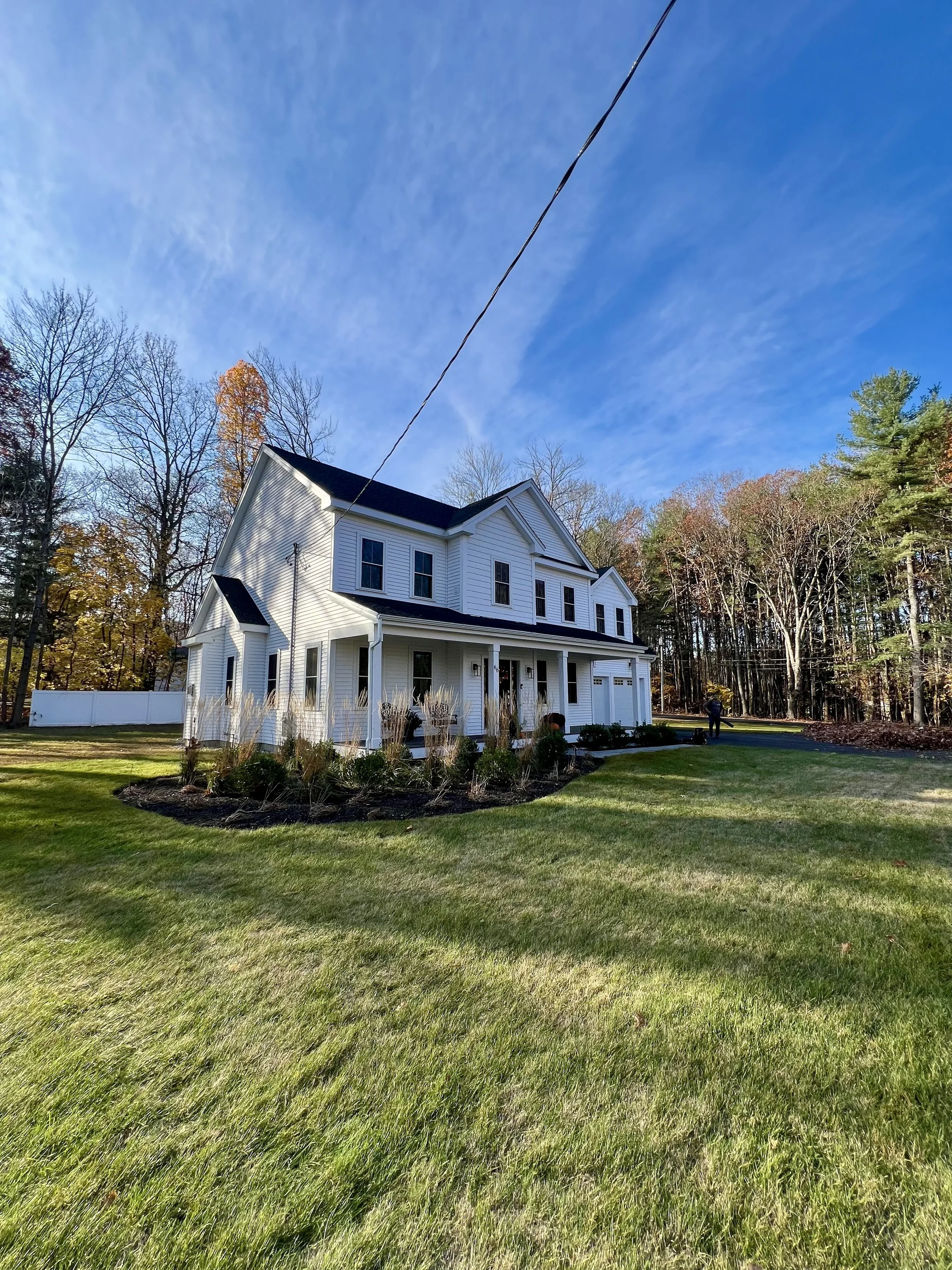 White two-story house with a front porch, black roof, and multiple windows, surrounded by a well-maintained lawn and trees with autumn leaves, under a clear blue sky.