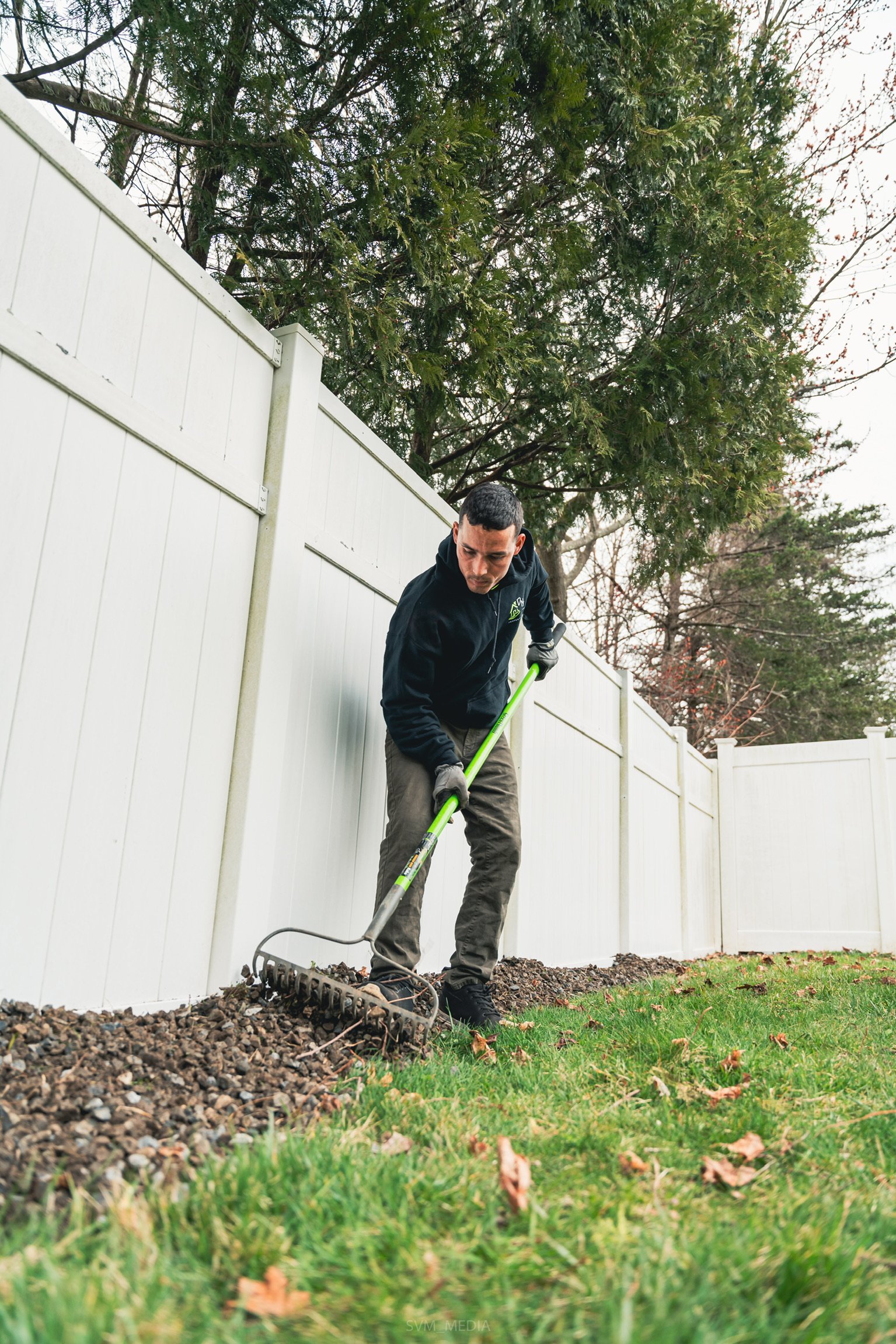 A man using a rake to level small rocks along the edge of a lawn, next to a white fence and tall trees.
