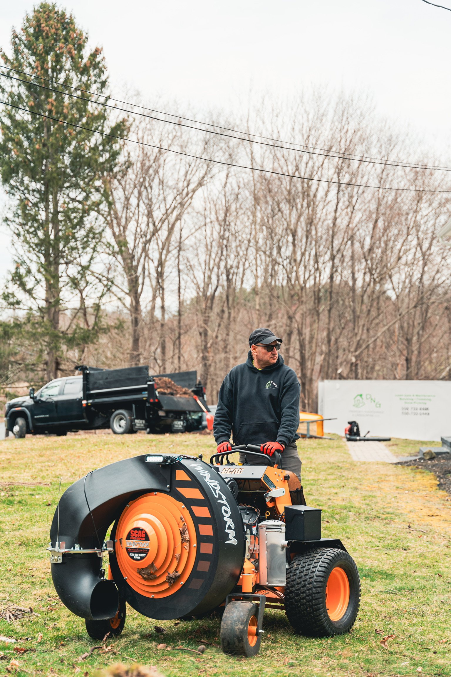 A man in a black hoodie, red gloves, and sunglasses operates a leaf blower machine on a lawn with trees and utility trucks in the background.
