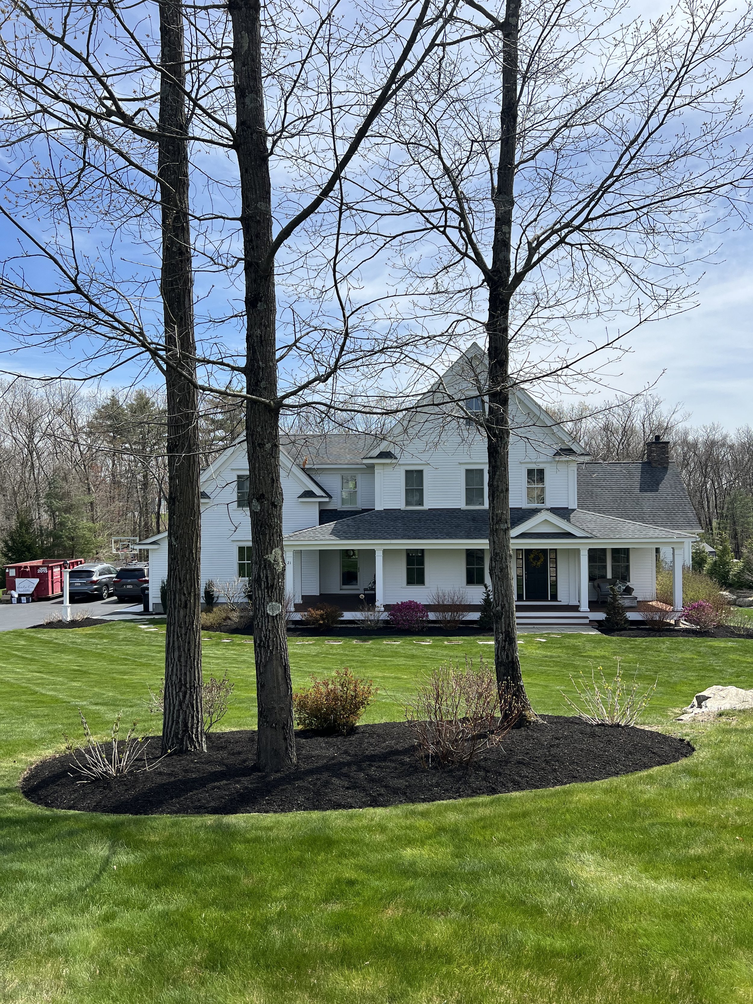 White two-story house with a dark roof, surrounded by a well-maintained green lawn and a landscaped flower bed with small shrubs and three large trees in the foreground.