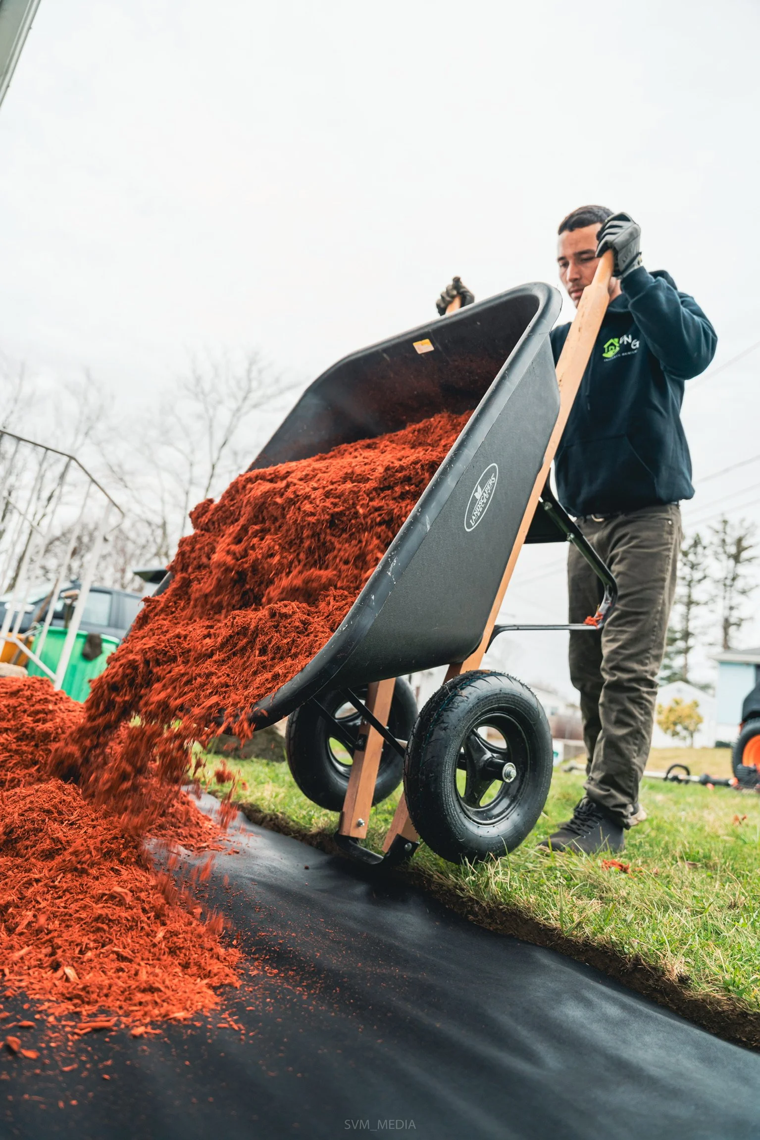 A person using a wheelbarrow to transport reddish mulch on a cloudy day in a yard.