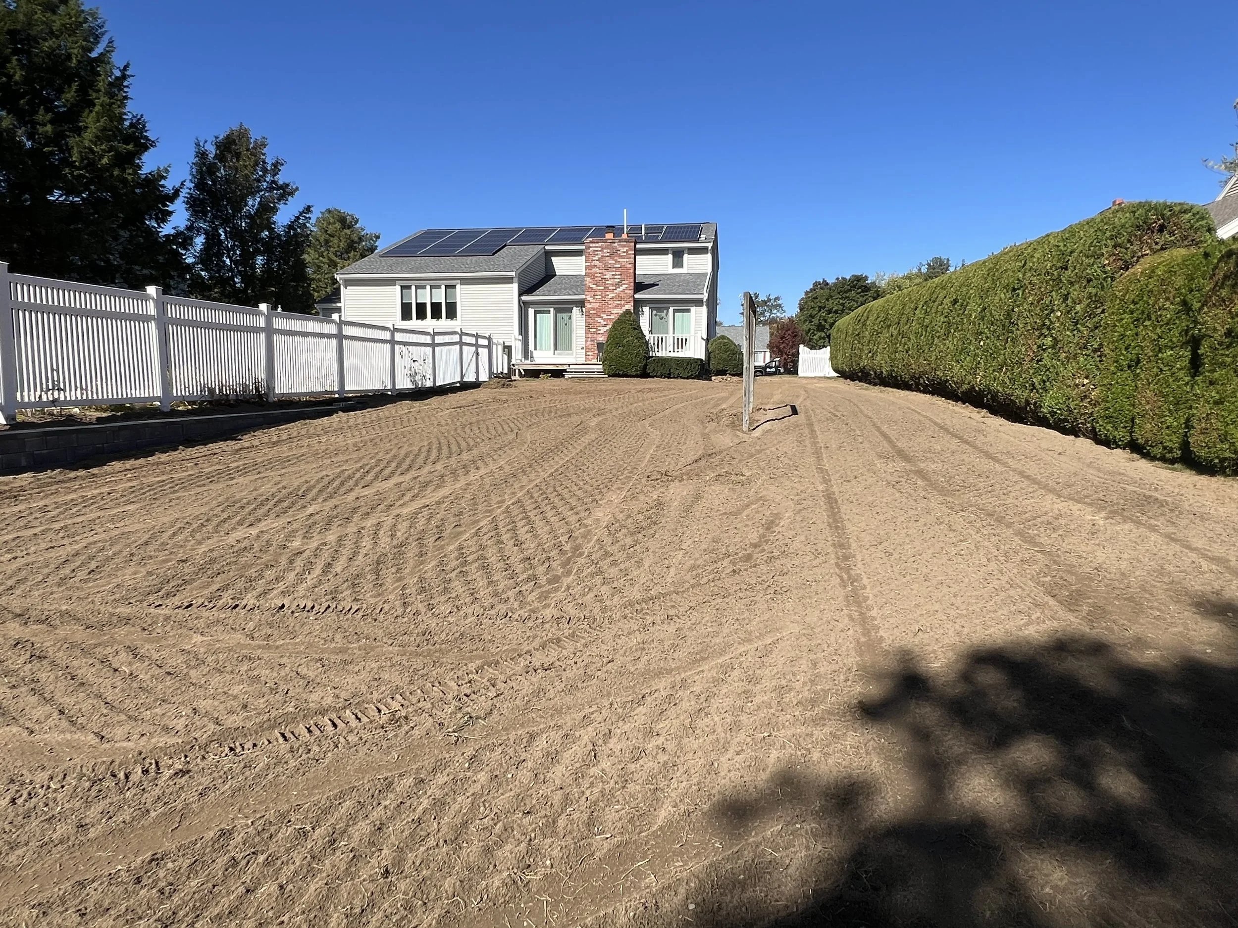 Large backyard with prepared dirt surface, white fence on the left, tall hedges on the right, and a house with solar panels on the roof in the background.