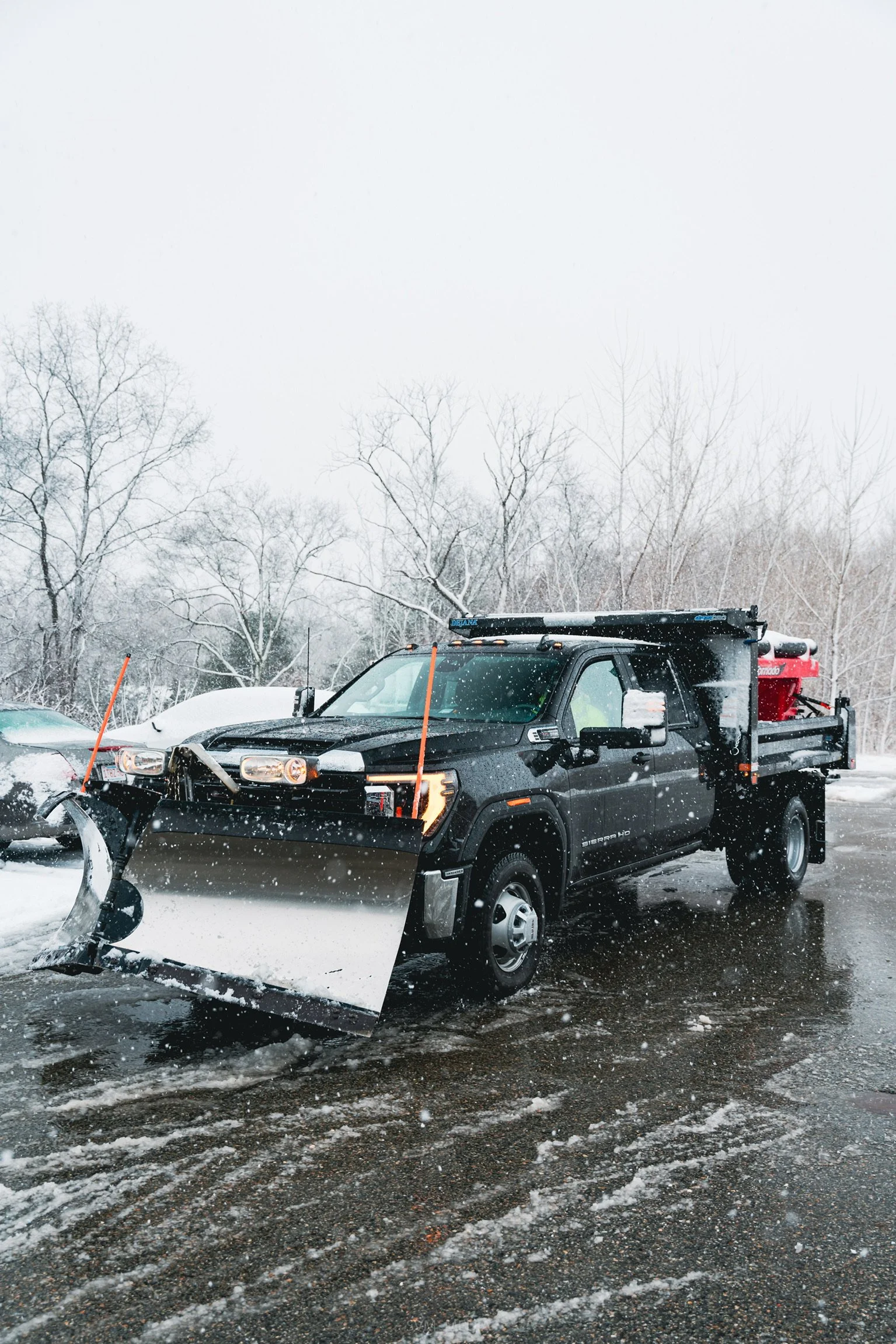 A black snow plow truck clearing snow from a parking lot on a snowy day, with leafless trees in the background.