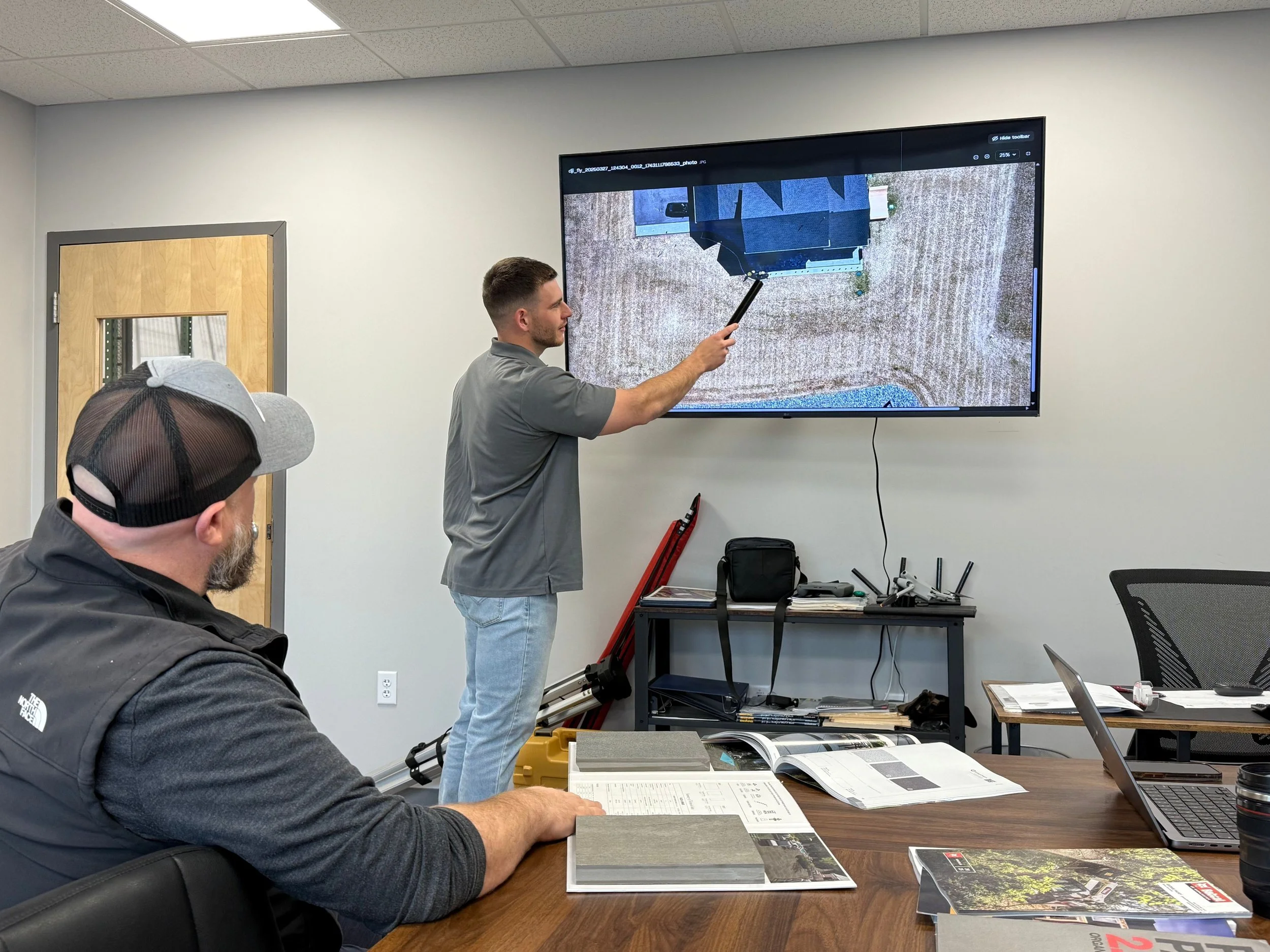 Man presenting an aerial construction site image on a wall-mounted monitor during an office meeting