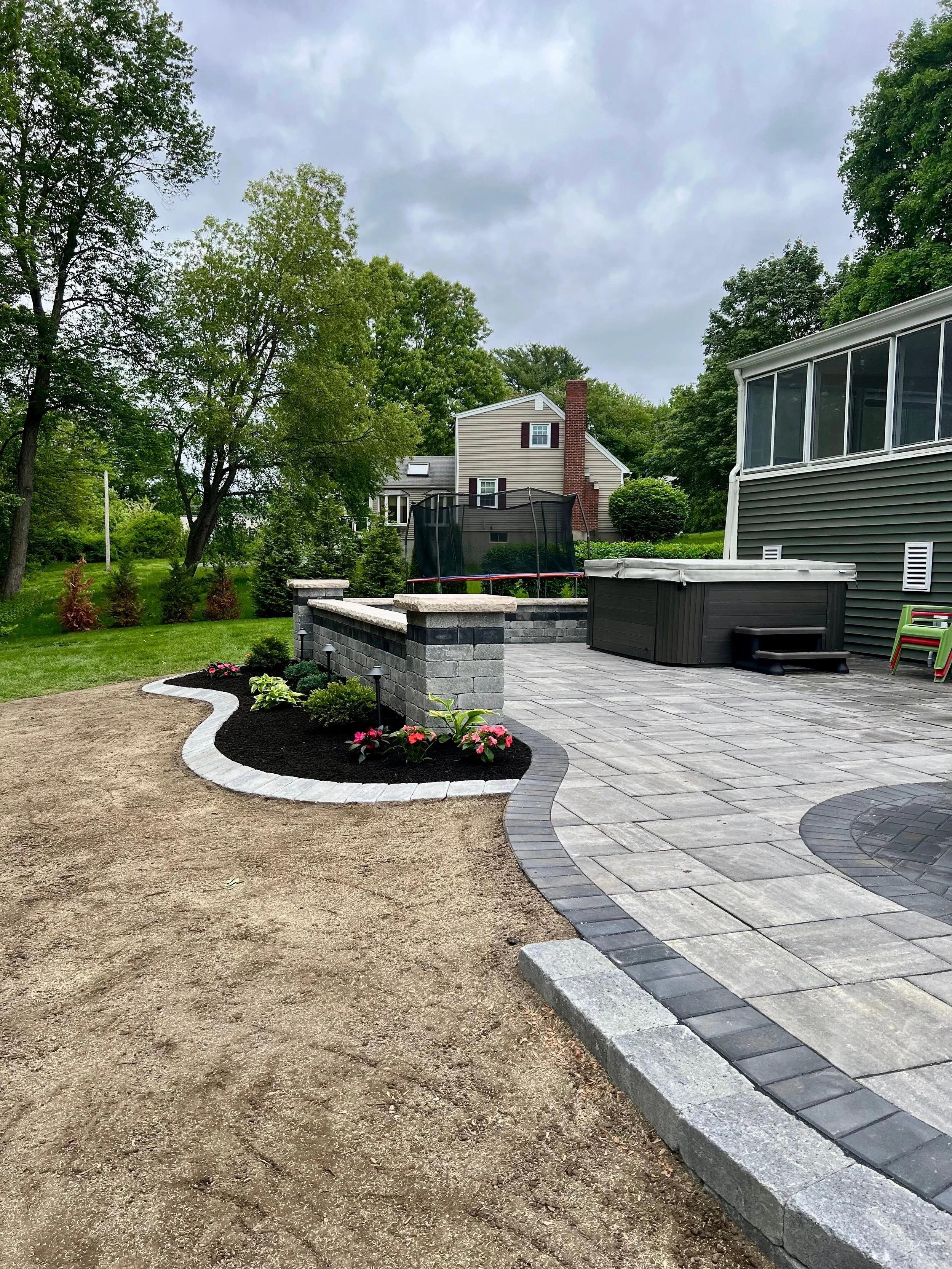 A landscaped backyard with a stone patio, a hot tub, a trampoline, and a garden bed with flowers, surrounded by green grass and trees under a cloudy sky.