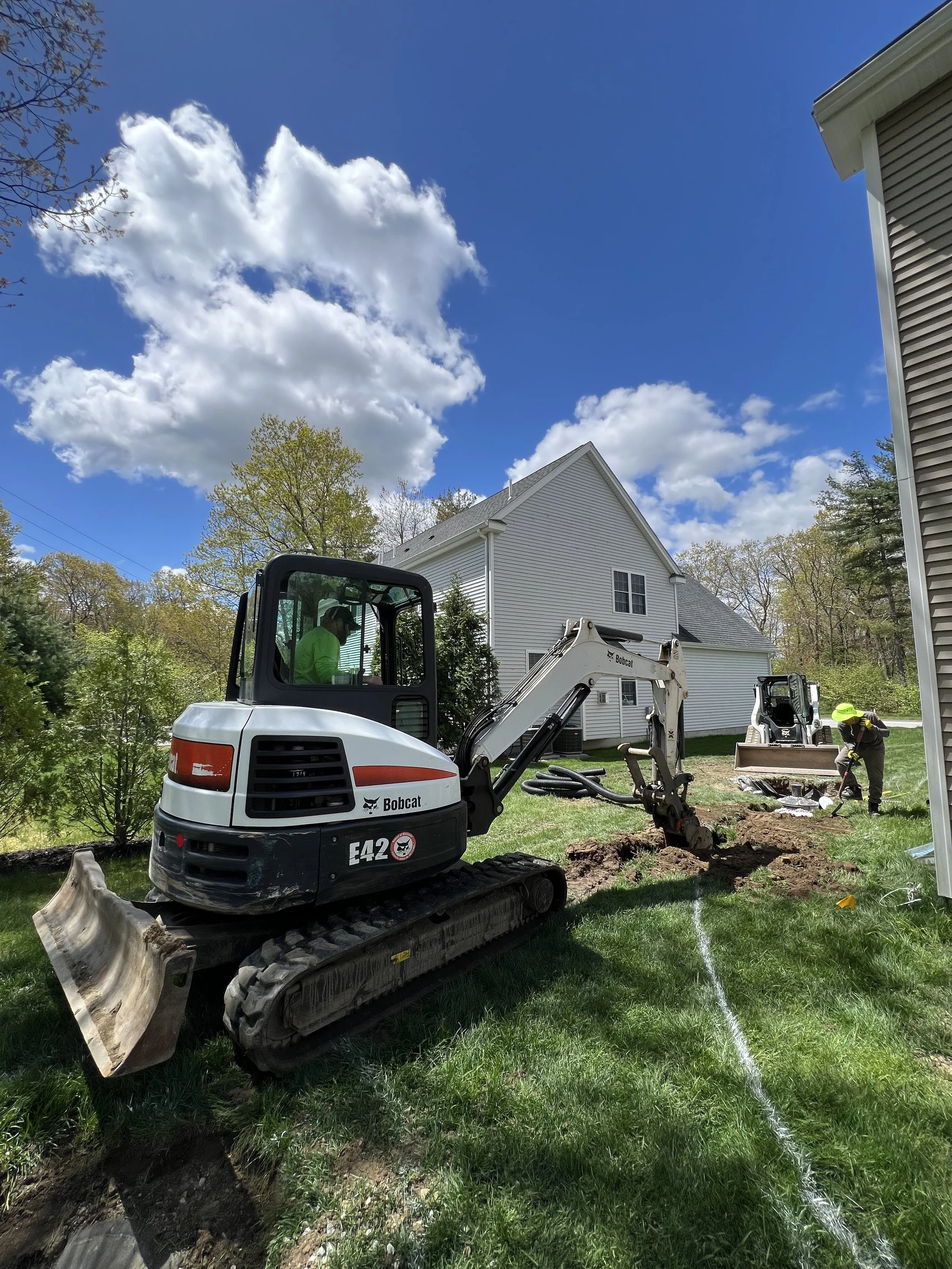 Bobcat excavator and workers digging a trench beside a house on a sunny day