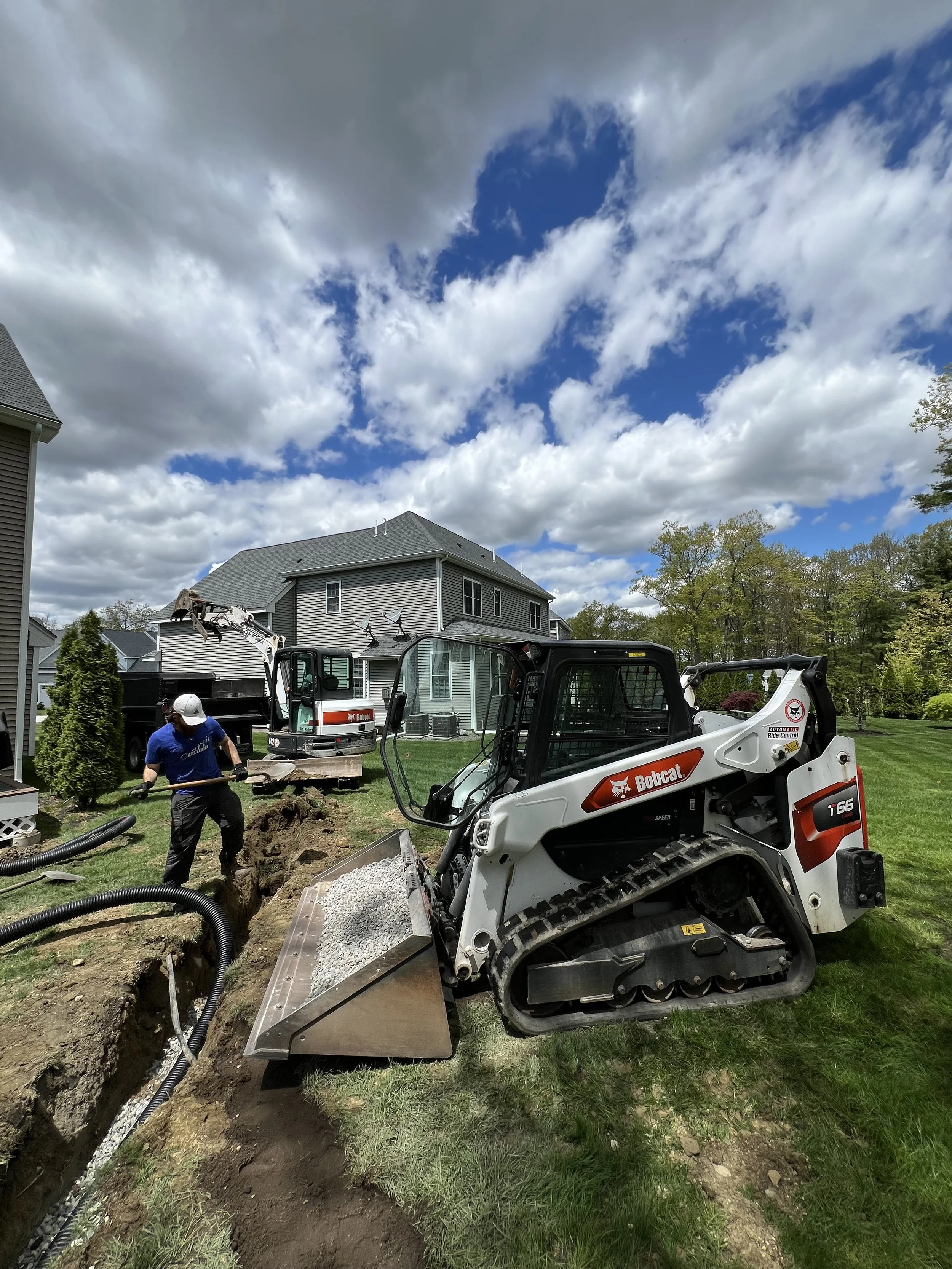 Bobcat skid-steer loaders and a worker excavating a trench in a suburban yard beside a house.