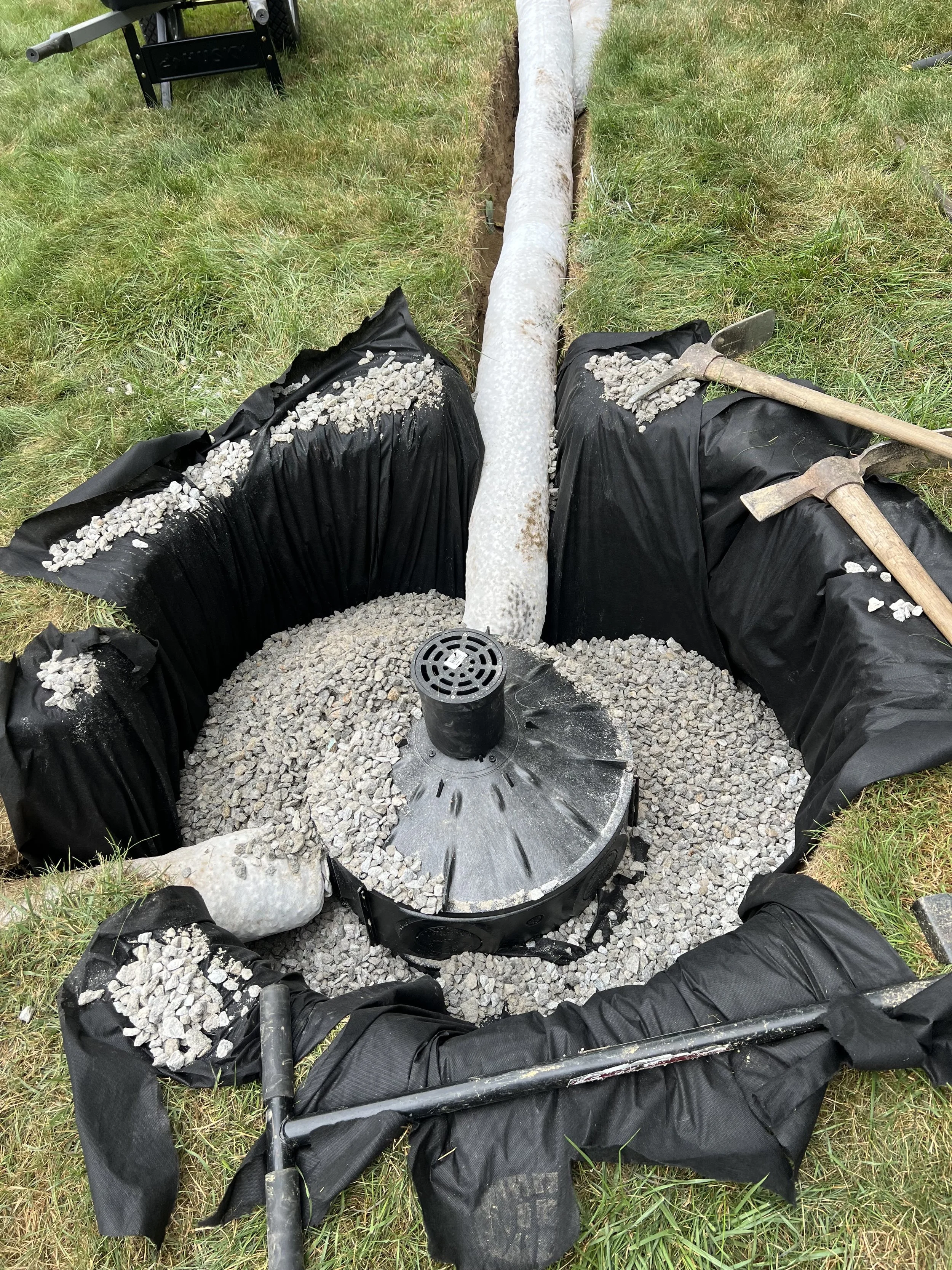 Backyard pond installation with a large liner, gravel, drain, pipe, and hand tools in a grassy yard
