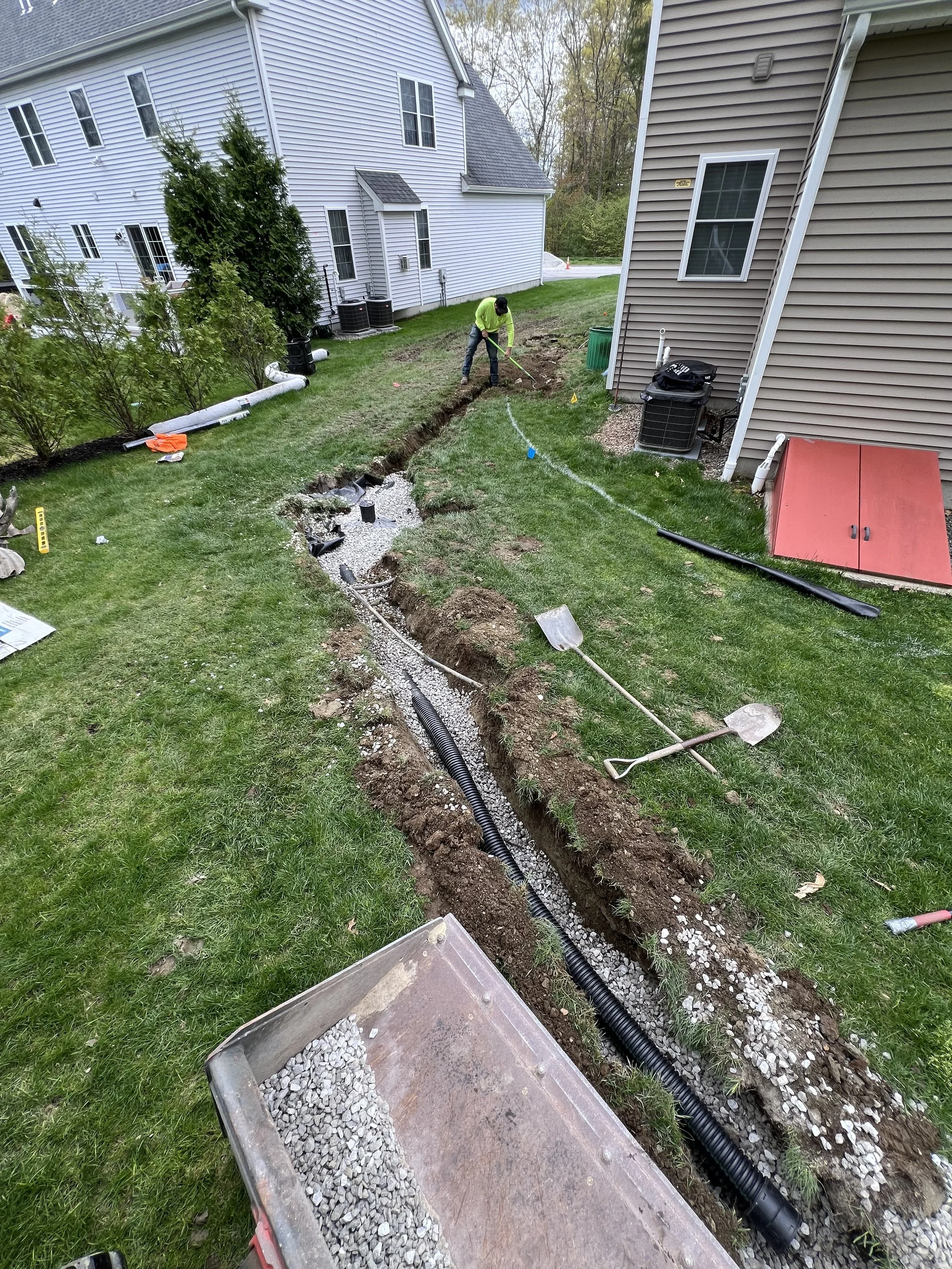 Worker digging a drainage trench for corrugated pipe between suburban homes, with gravel, shovels, and landscaping equipment.