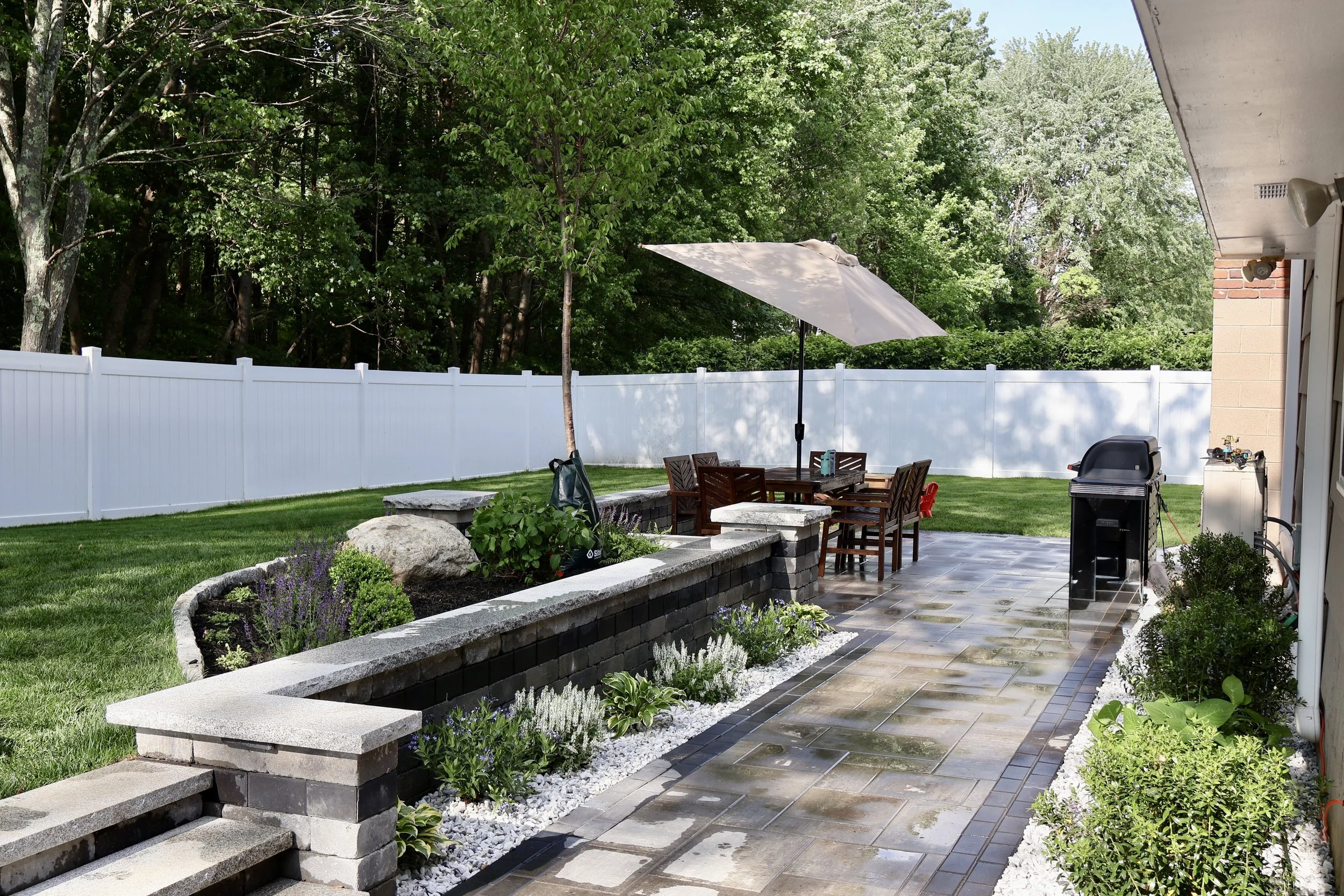 Landscaped backyard patio with dining table, umbrella, grill, stone retaining walls, gardens, lawn, and white privacy fence
