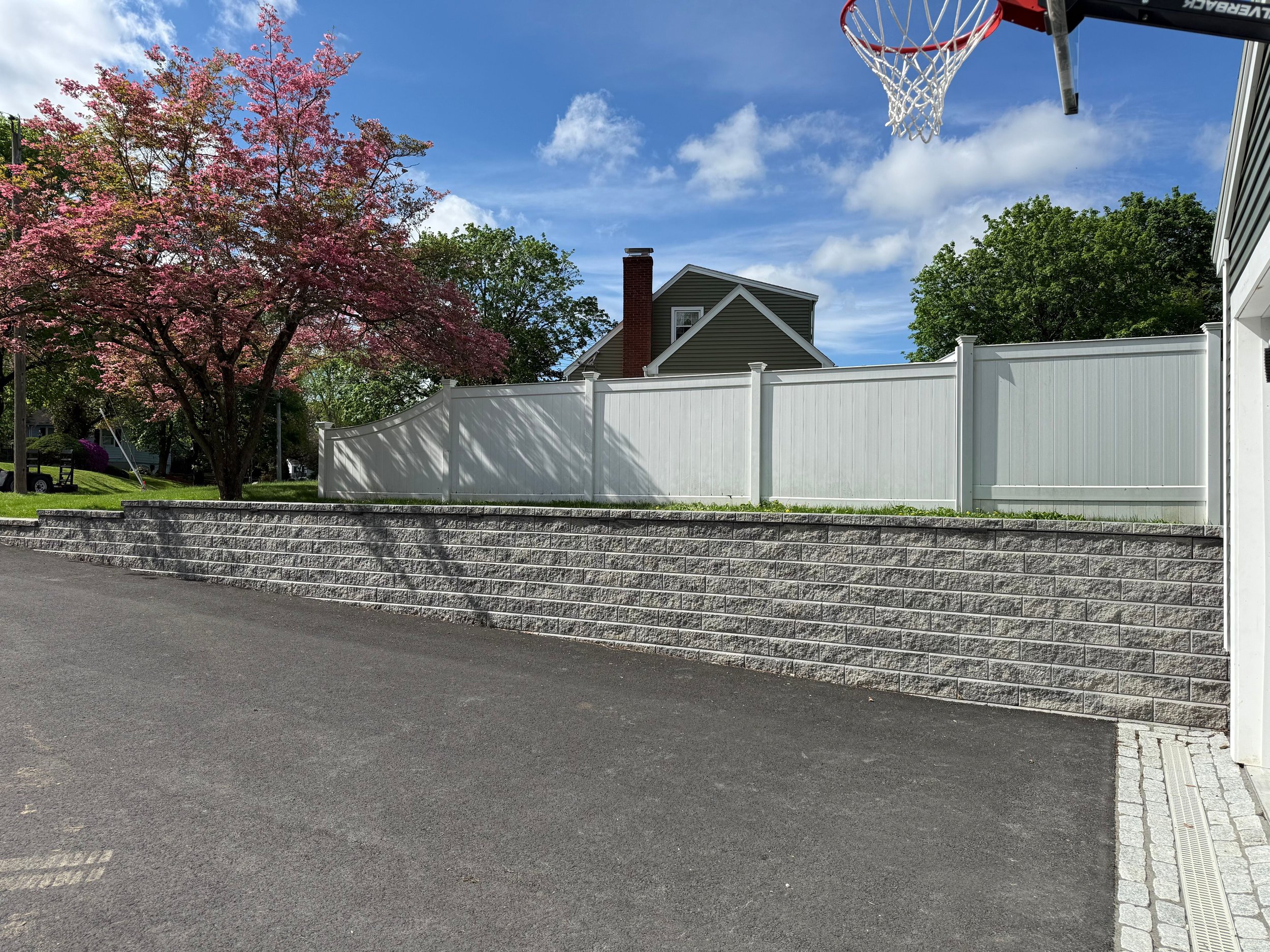 Residential backyard with a basketball hoop, a pink flowering tree, a white privacy fence, and a stone retaining wall along the street under a partly cloudy blue sky.