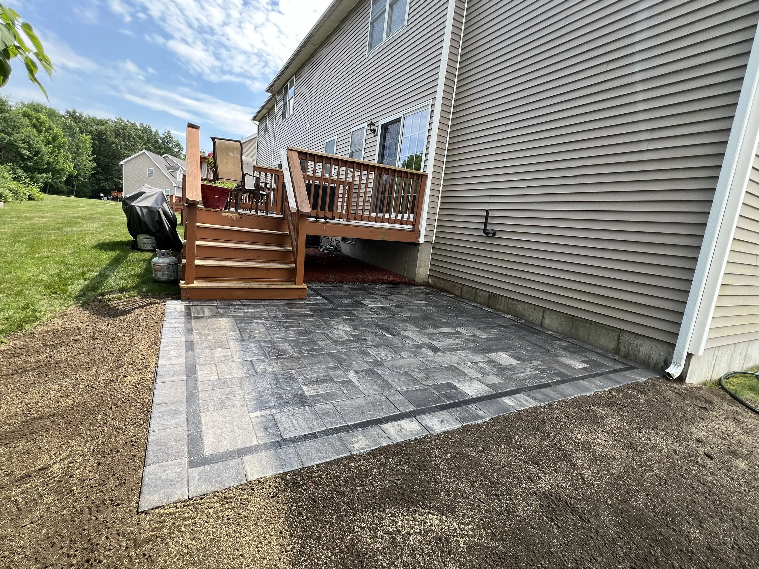 Backyard with newly paved patio, wooden deck stairs, beige house siding, and neighboring houses in the background under a partly cloudy sky.