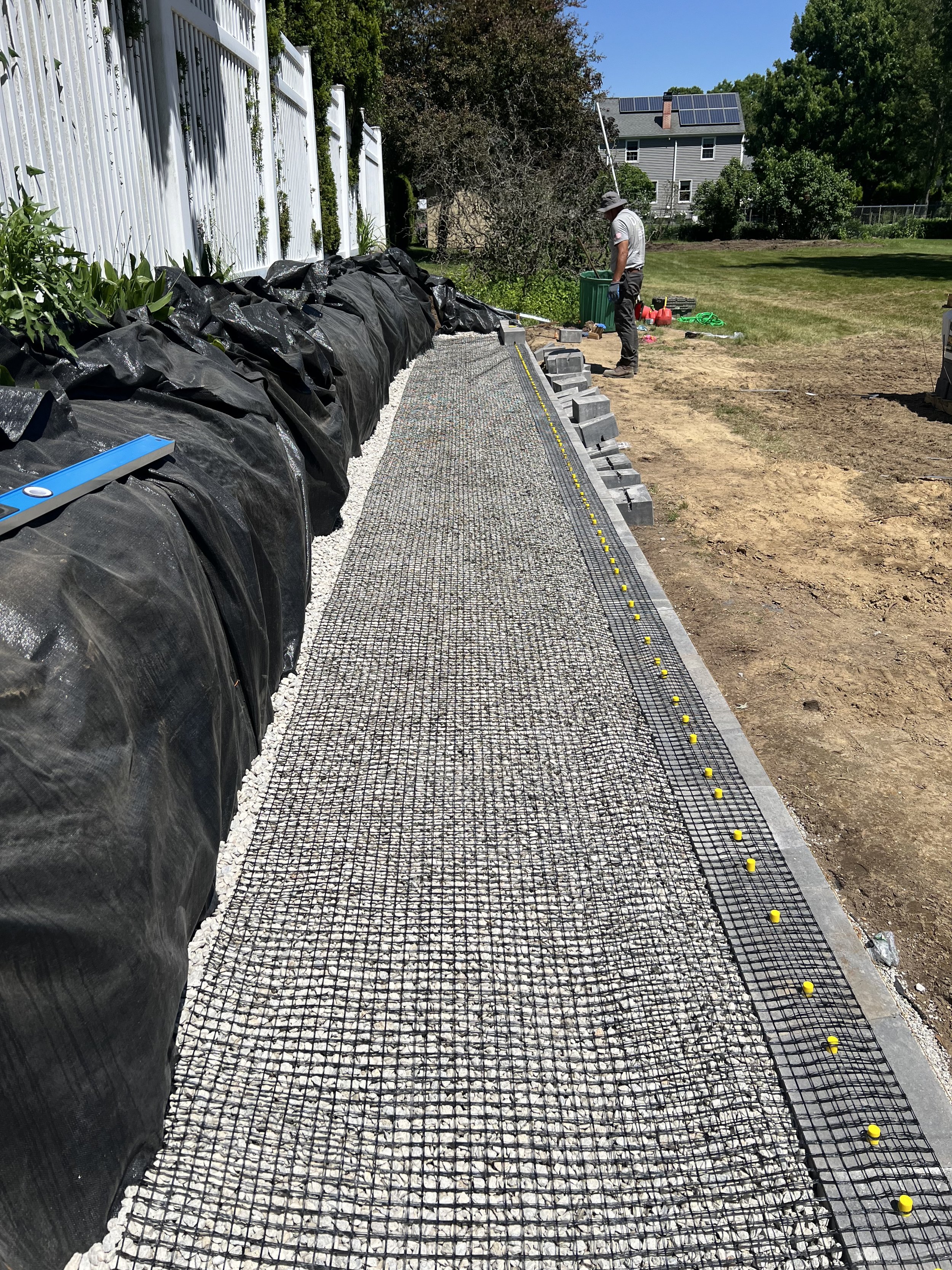 A backyard garden area under construction with a gravel path being laid, black plastic sheeting on one side, and a man working nearby. There is a white fence, green trees, and a house with solar panels in the background.