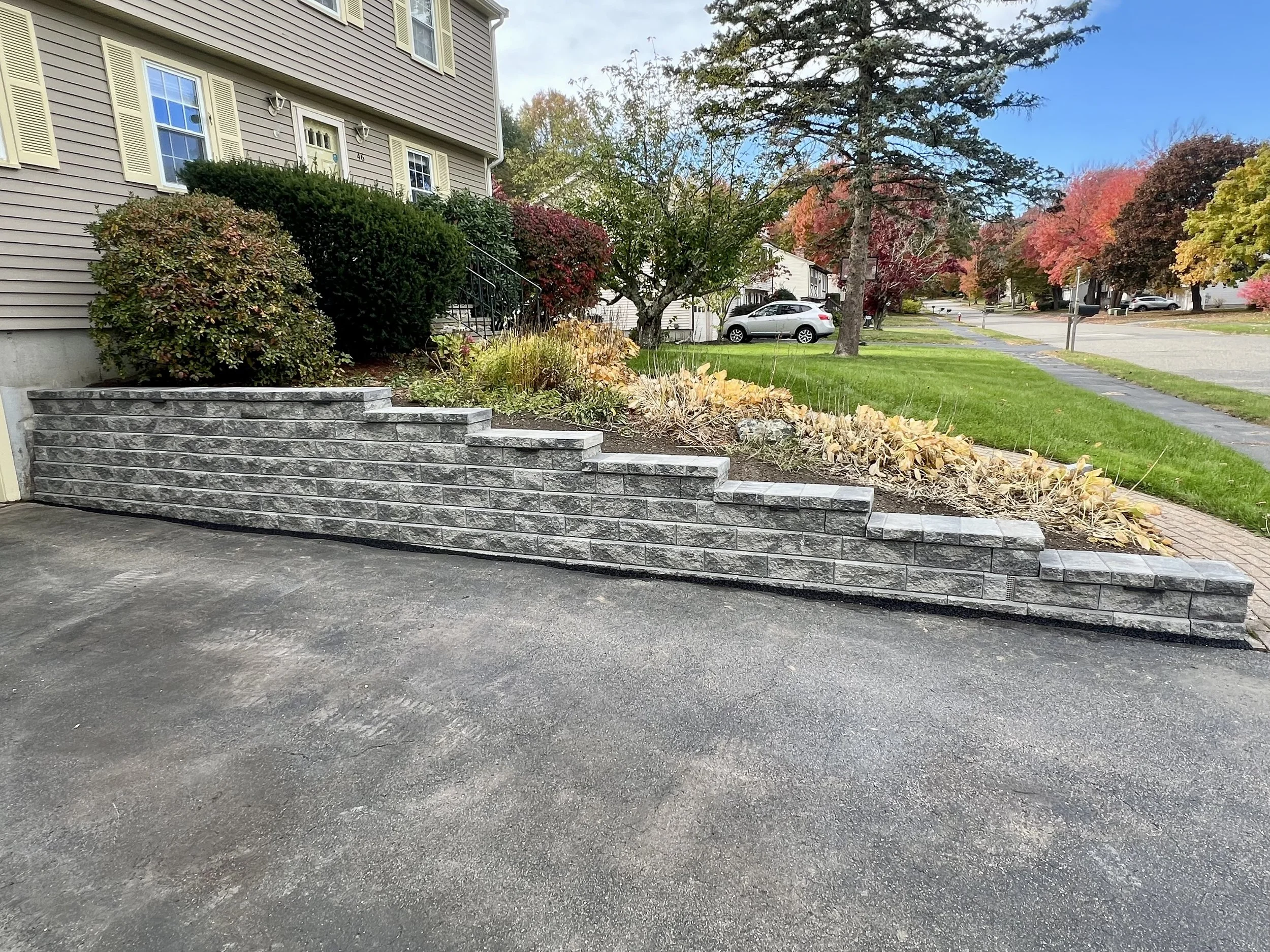 Stone retaining wall with steps, adjacent to a driveway, in front of a house with shrubs and autumn trees.