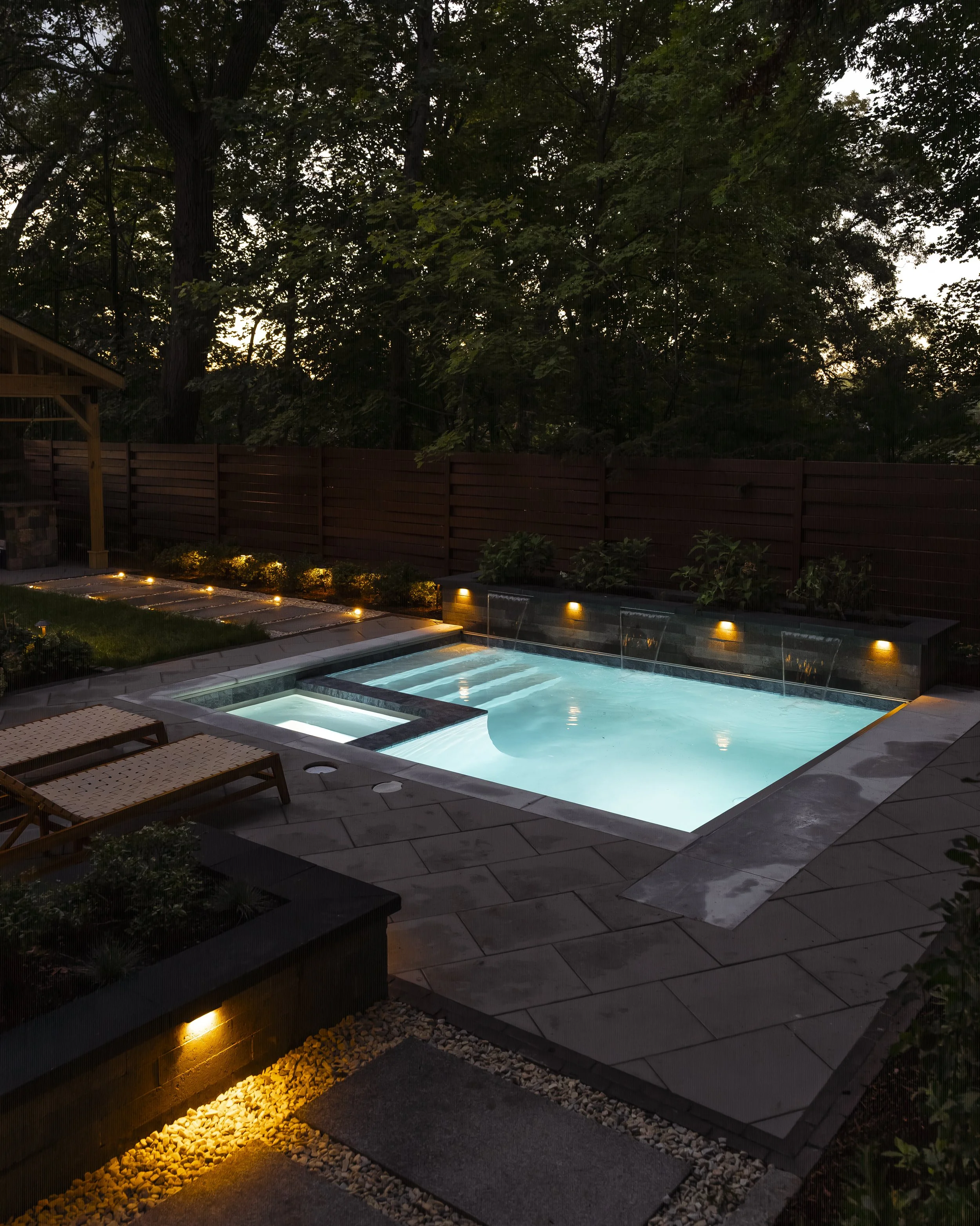Illuminated backyard swimming pool with water features, hot tub, lounge chairs, and wooded privacy fence at dusk