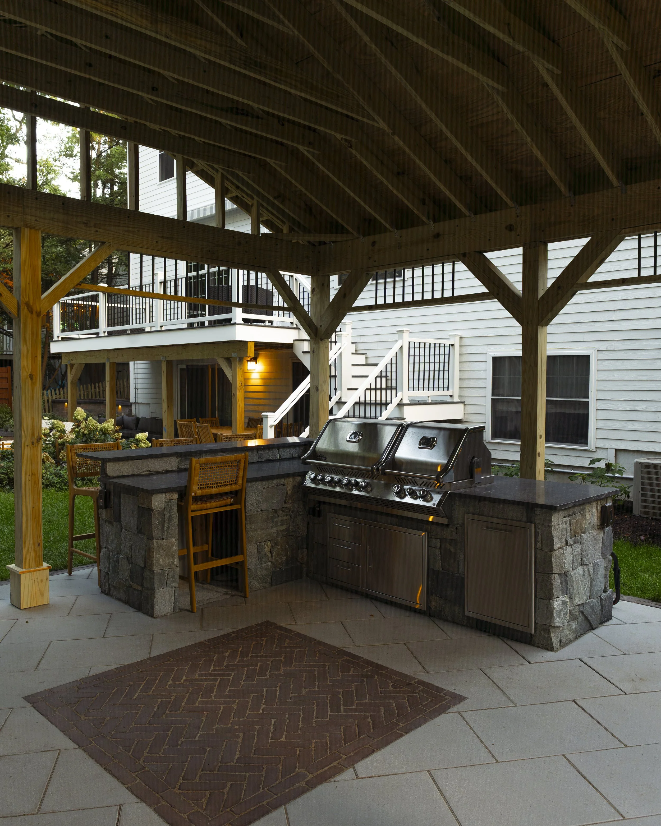 Covered outdoor patio with a stainless steel grill, dining furniture, stone counters, and wooden pergola beside a house