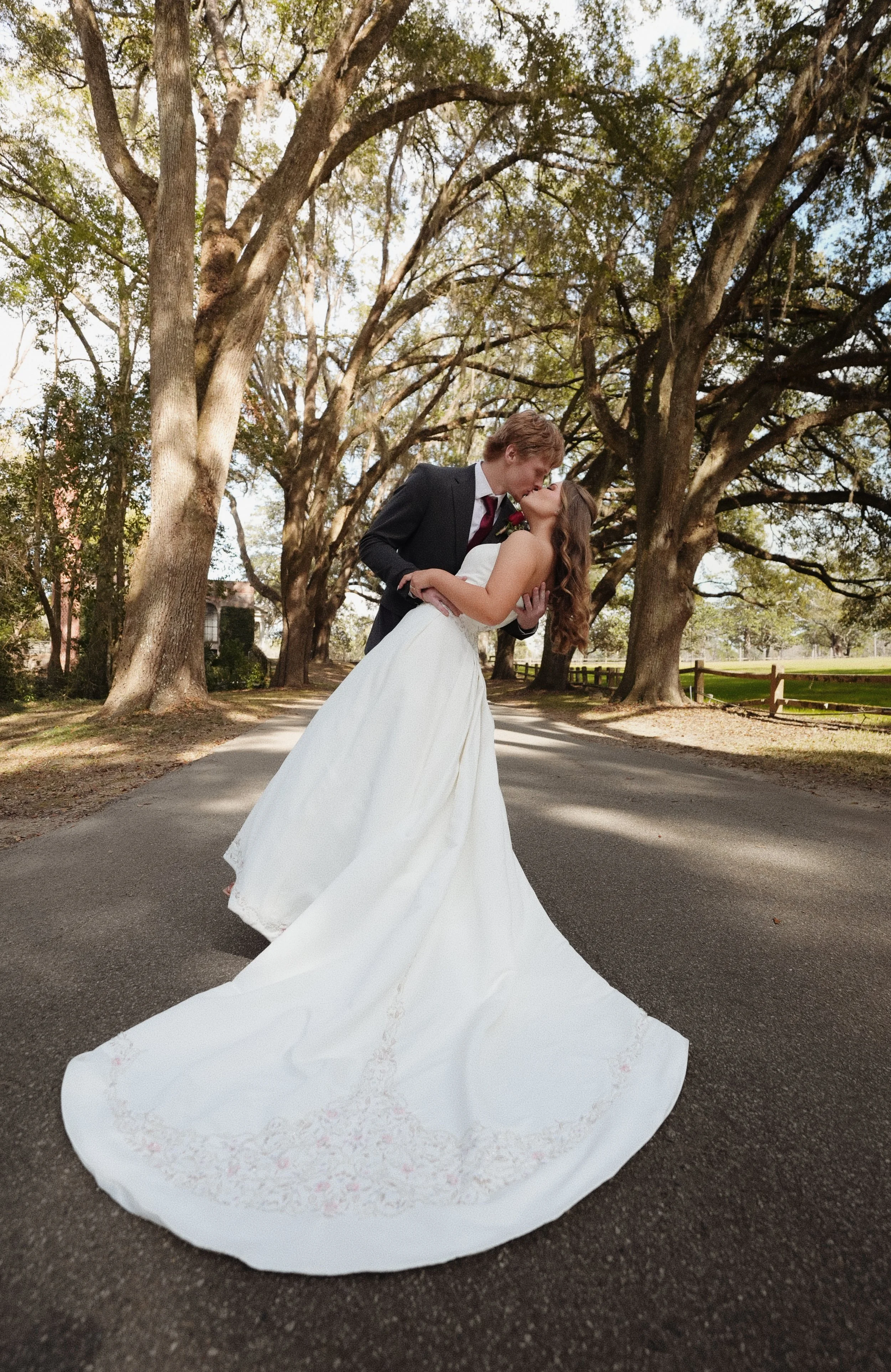 A newlywed couple is kissing under a large canopy of tall trees on a paved path, with the bride wearing a white gown and the groom in a black suit, in a scenic outdoor setting.