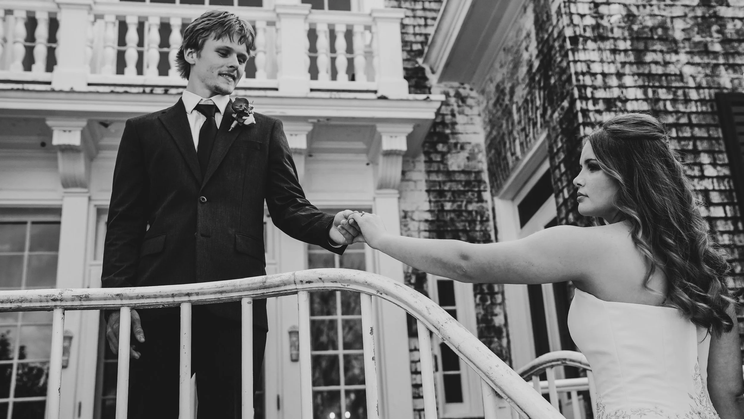 A black-and-white photograph of a man and a woman on a balcony. The man, wearing a suit with a boutonnière, is holding hands with the woman. The woman is wearing a strapless dress with wavy hair, and they are posing in front of a large house with a b