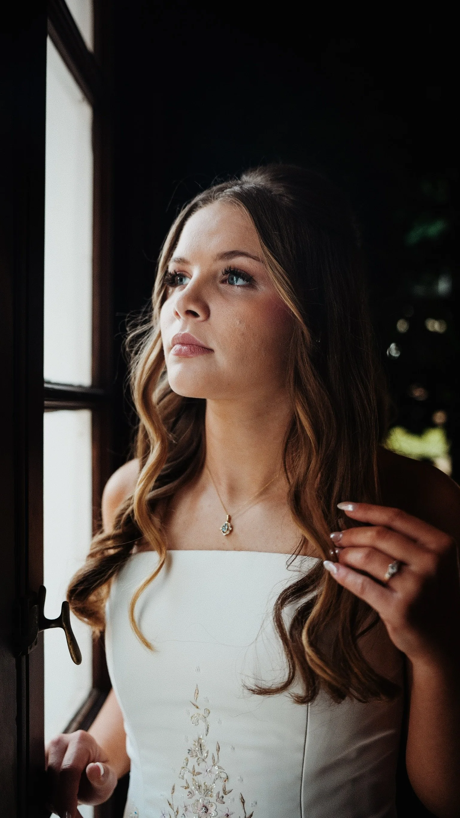 Young woman with long wavy brown hair, wearing a white floral dress, jewelry, and makeup, looking out a window.