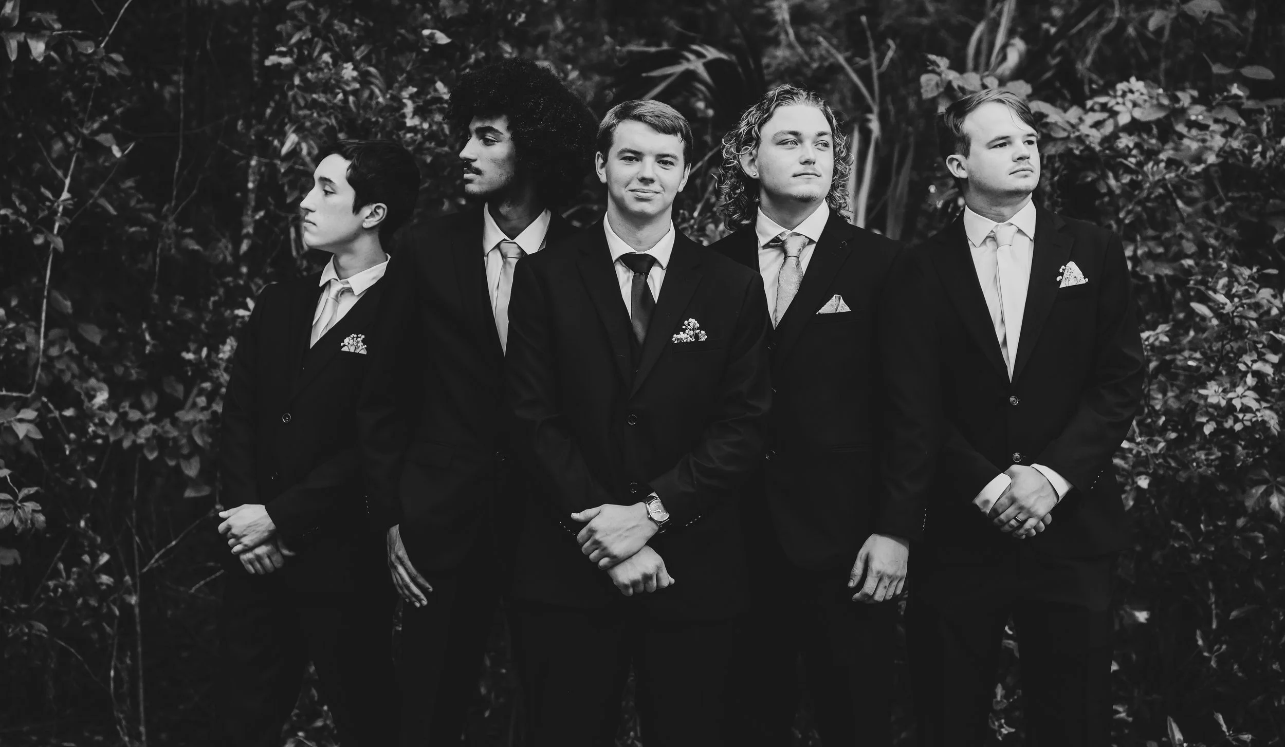 Five young men in suits stand in a line outdoors, looking in different directions, with a background of dense foliage.