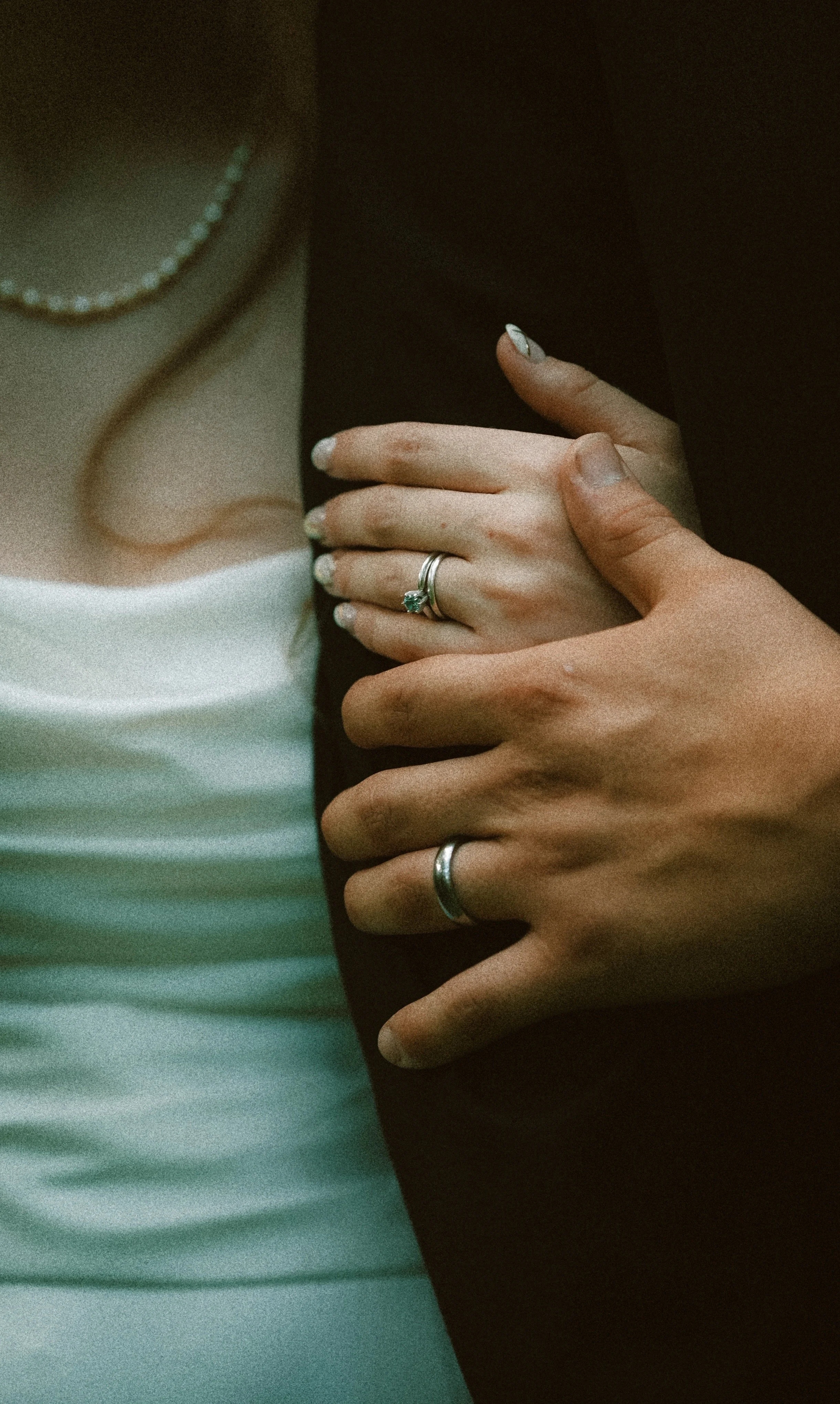 Close-up of two hands, one dark-skinned and one light-skinned, touching each other gently. Both hands are wearing rings, and part of a white shirt, a dark jacket, and a pearl necklace are visible.
