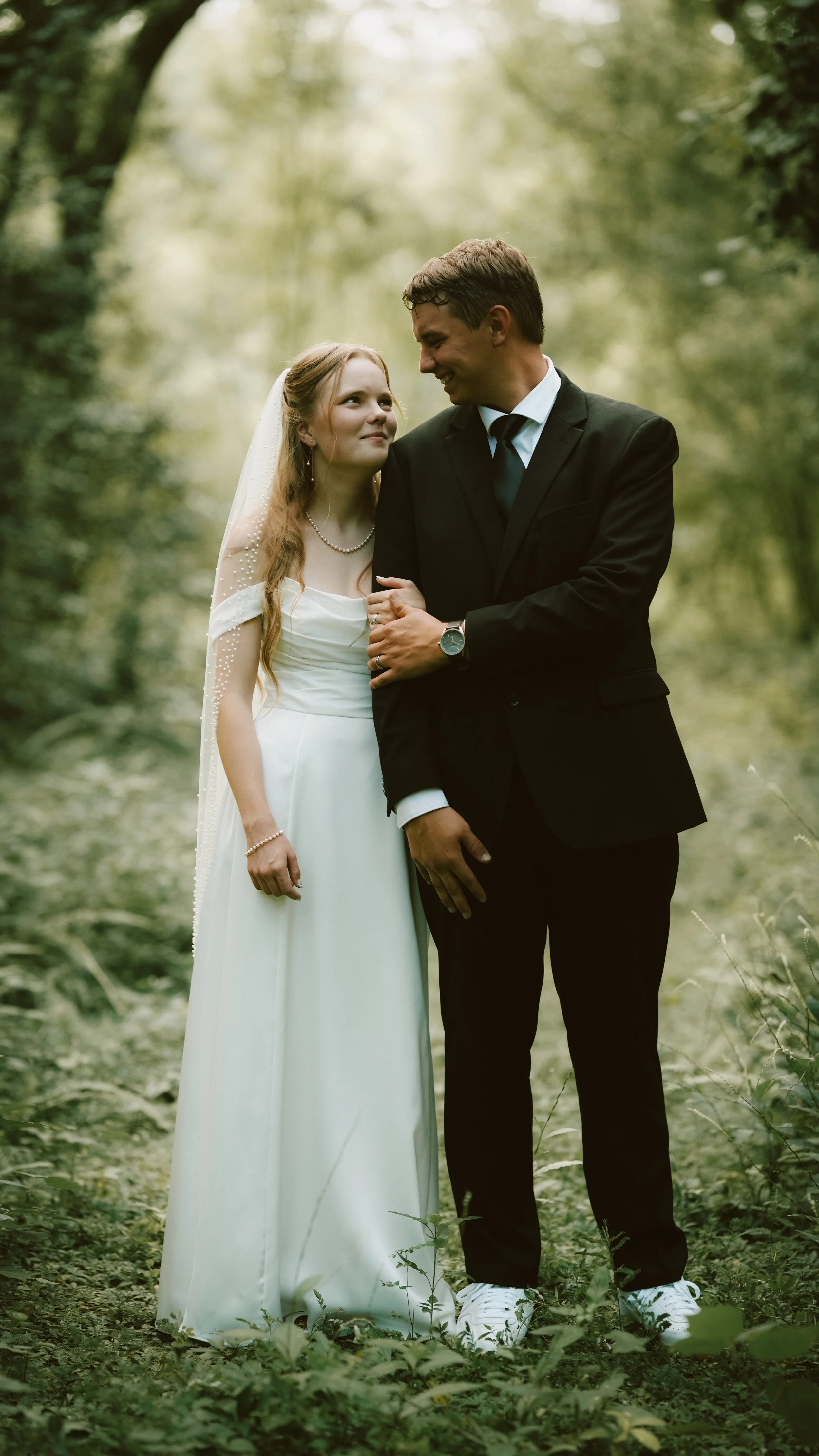 A bride and groom standing together in a lush forest, dressed in wedding attire, gazing into each other's eyes with smiles.