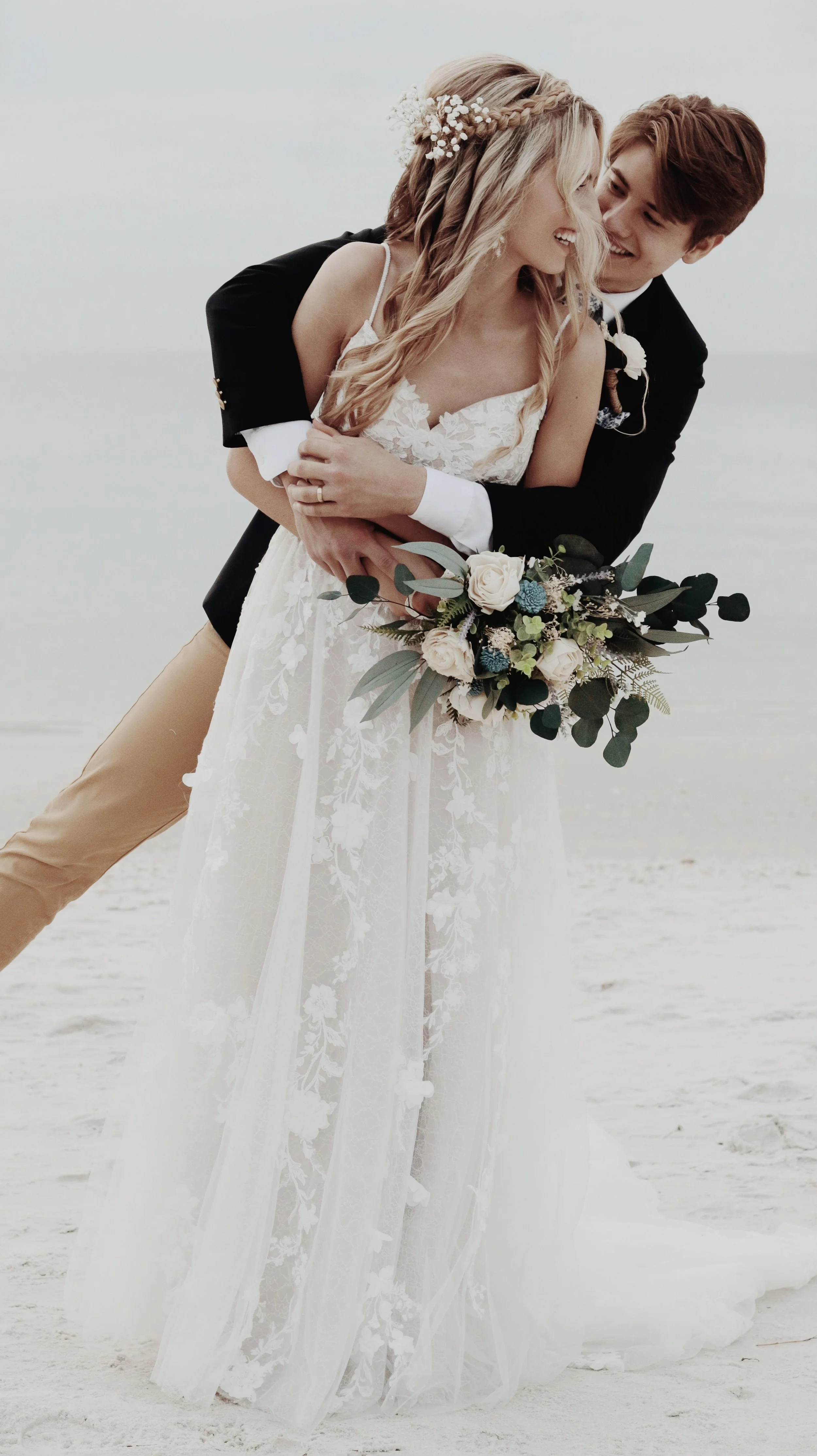 A happy couple at their wedding on the beach, with the groom embracing the bride from behind, holding her waist and smiling at her.