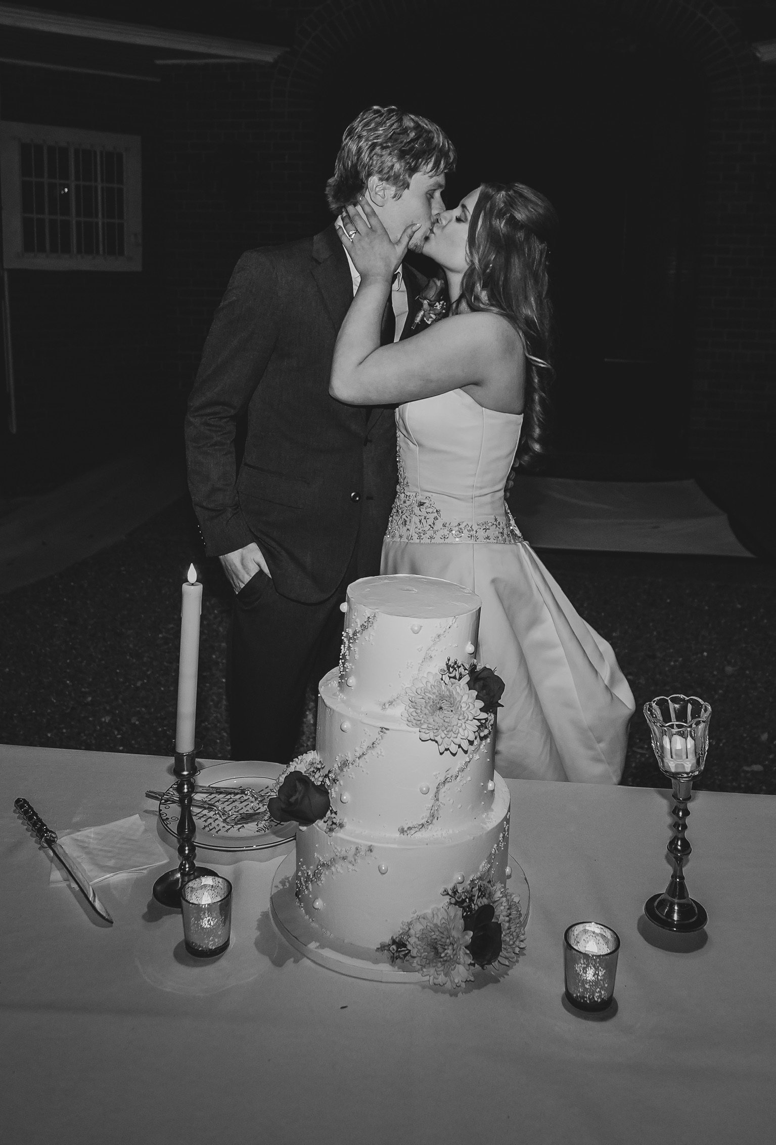 Black and white photo of a couple kissing at their wedding, with a wedding cake decorated with flowers on the table in front of them, and candles around it.