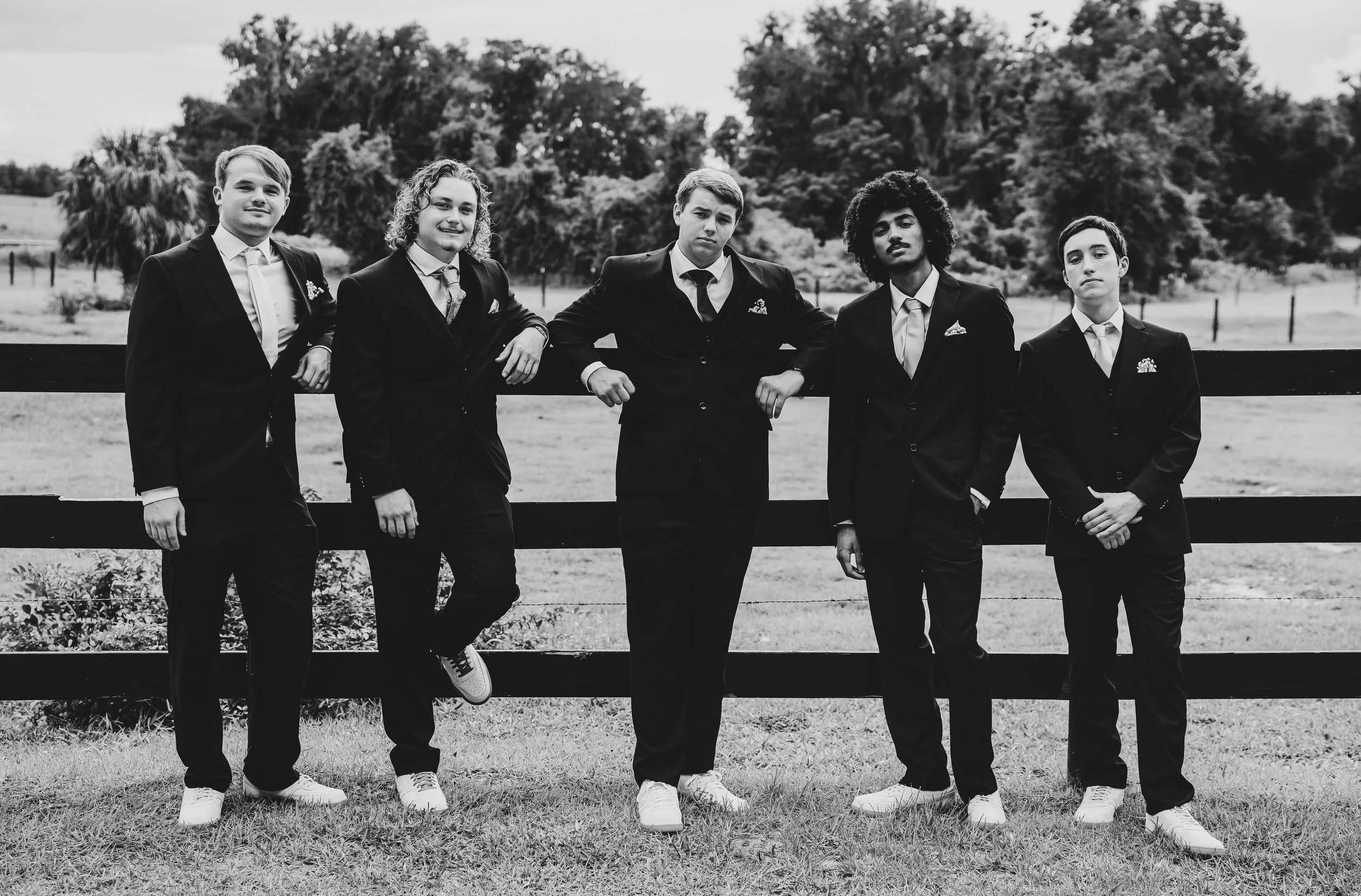 A black and white photo of five young men dressed in formal suits, standing outdoors with trees and a fence in the background.