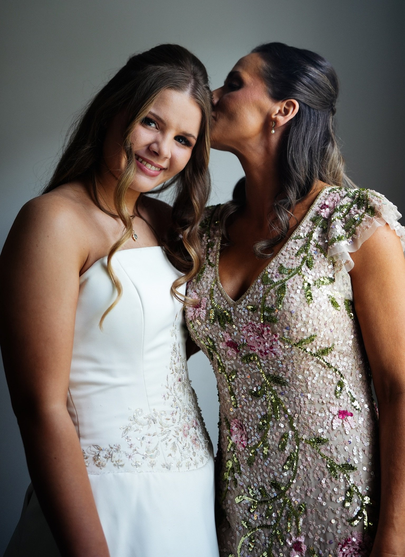 A young woman in a white dress smiling at the camera next to an older woman in a floral, sequined dress who is kissing her on the forehead.
