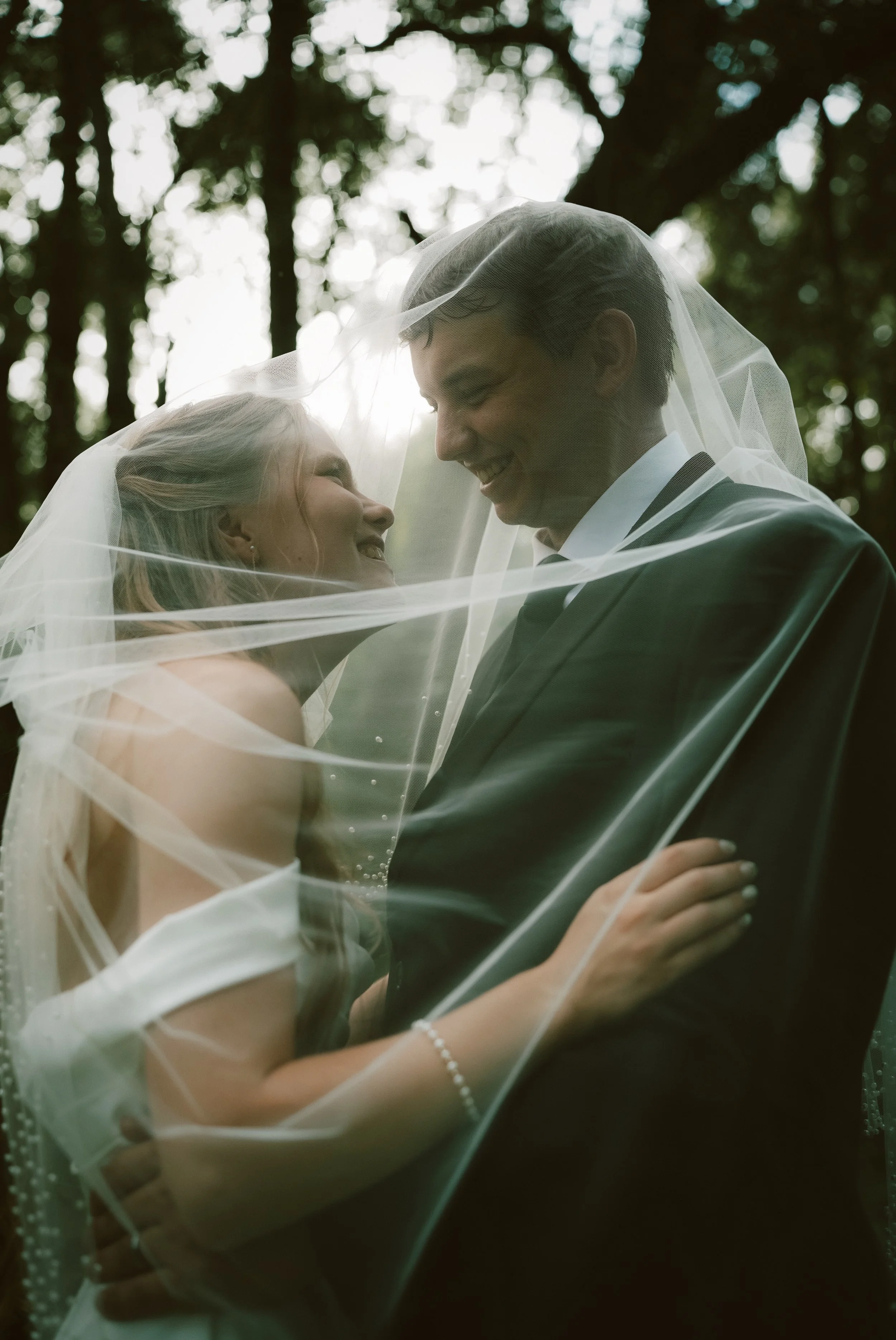 A bride and groom smiling and gazing at each other while standing close together outdoors, partially covered by a wedding veil. The background features blurred trees and natural light.
