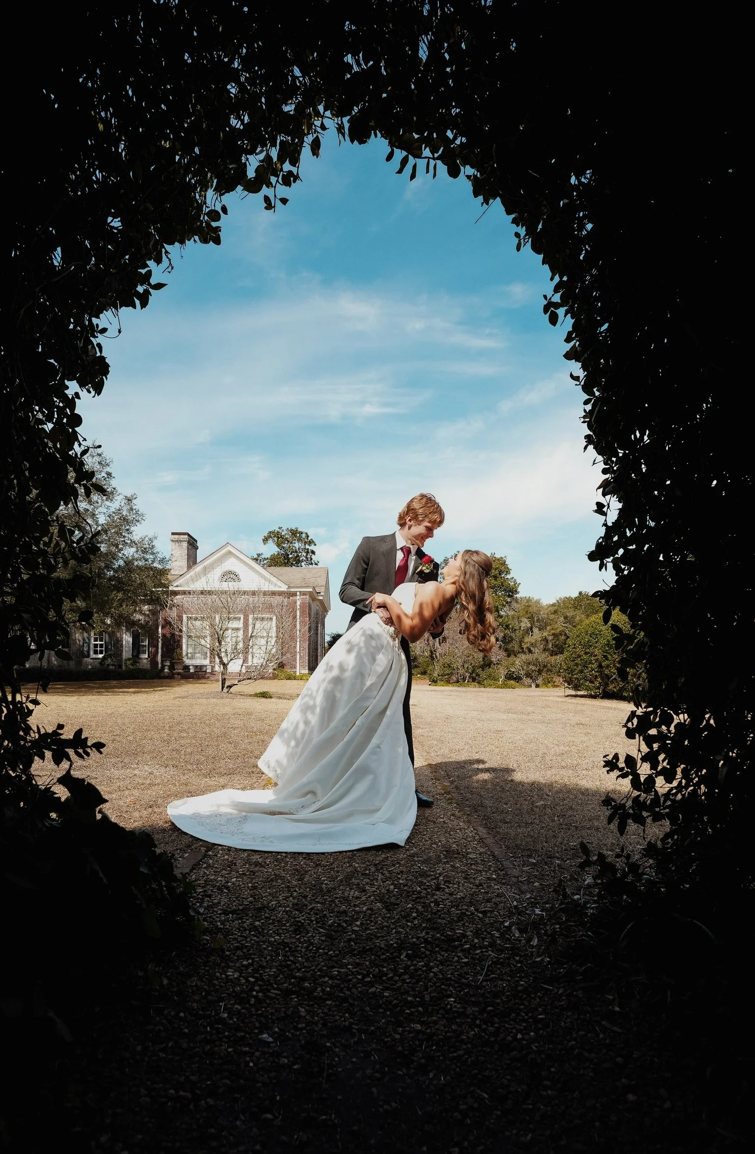 A couple dancing on the grass field outside a large historic white house, surrounded by trees, with a blue sky overhead, framed by dark foliage.