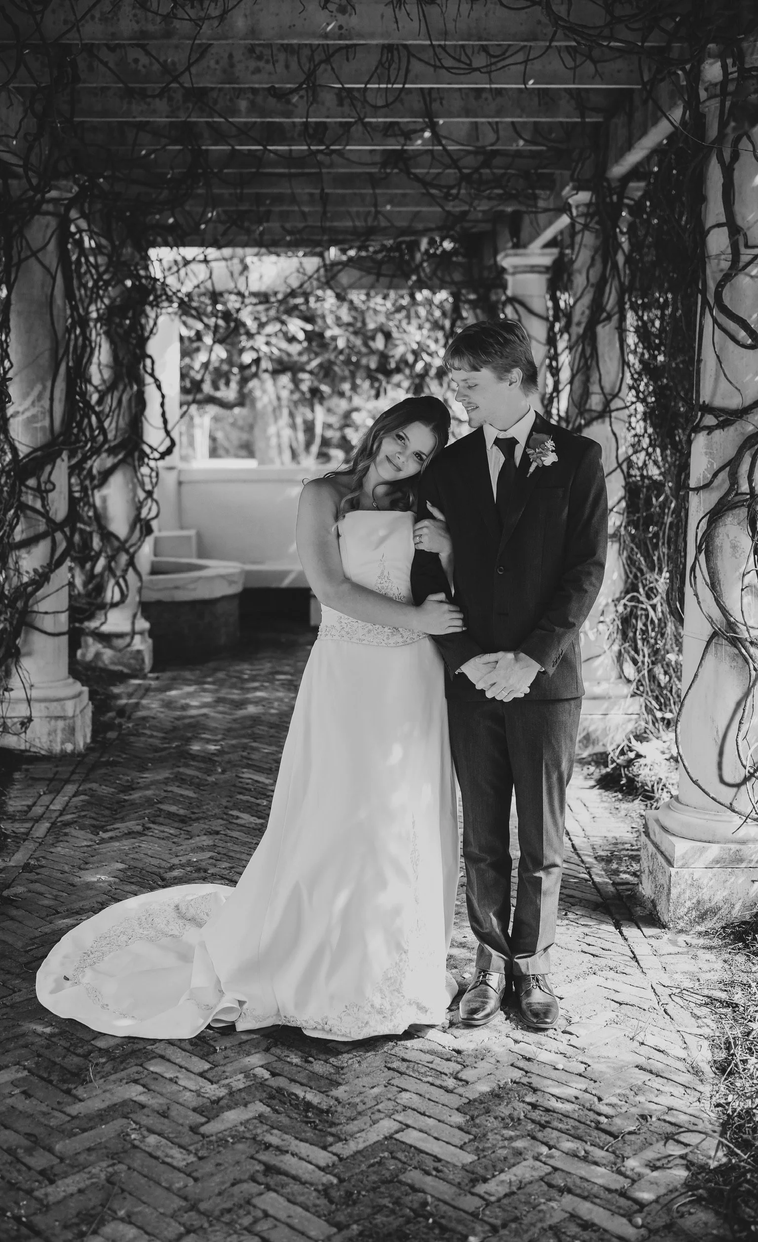 Black and white photo of a bride and groom standing under a pergola covered with vines. The bride wears a strapless wedding gown with a long train, and the groom wears a dark suit with a boutonnière. They are smiling and looking at each other.
