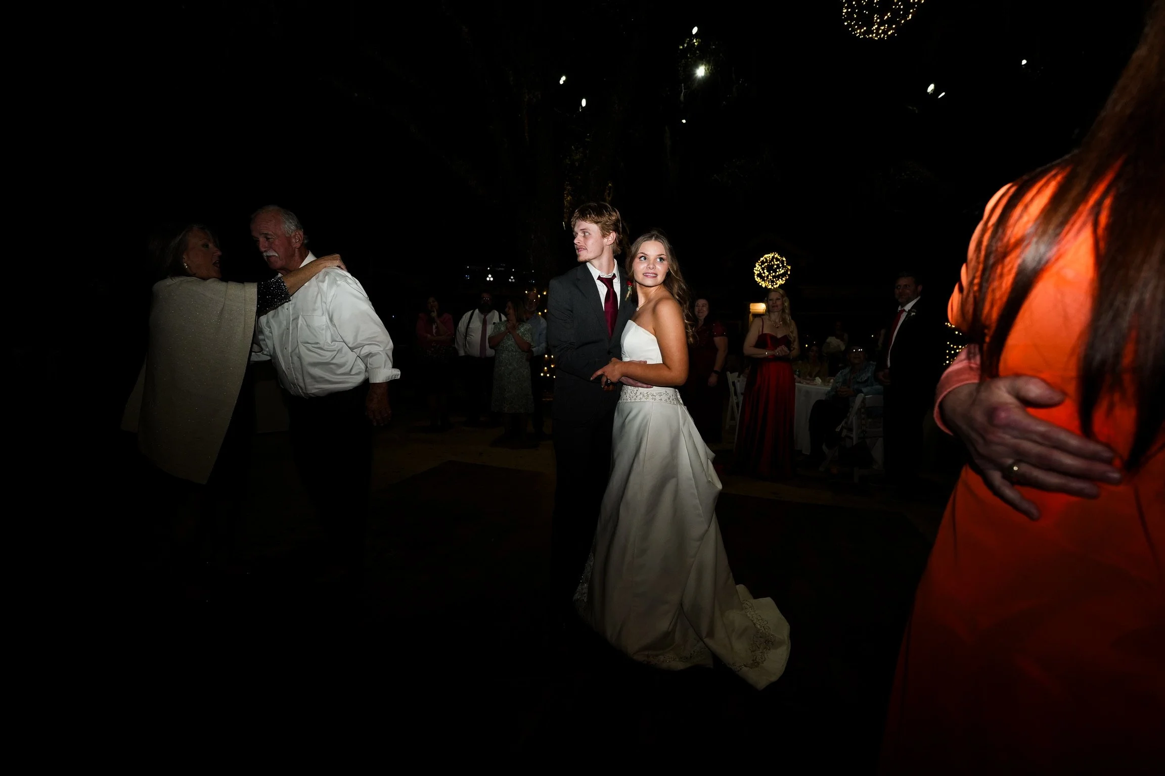 A bride and groom dance at their wedding reception under dark skies with fireworks above. The bride wears a white wedding gown, and the groom wears a dark suit. Guests can be seen in the background, celebrating.